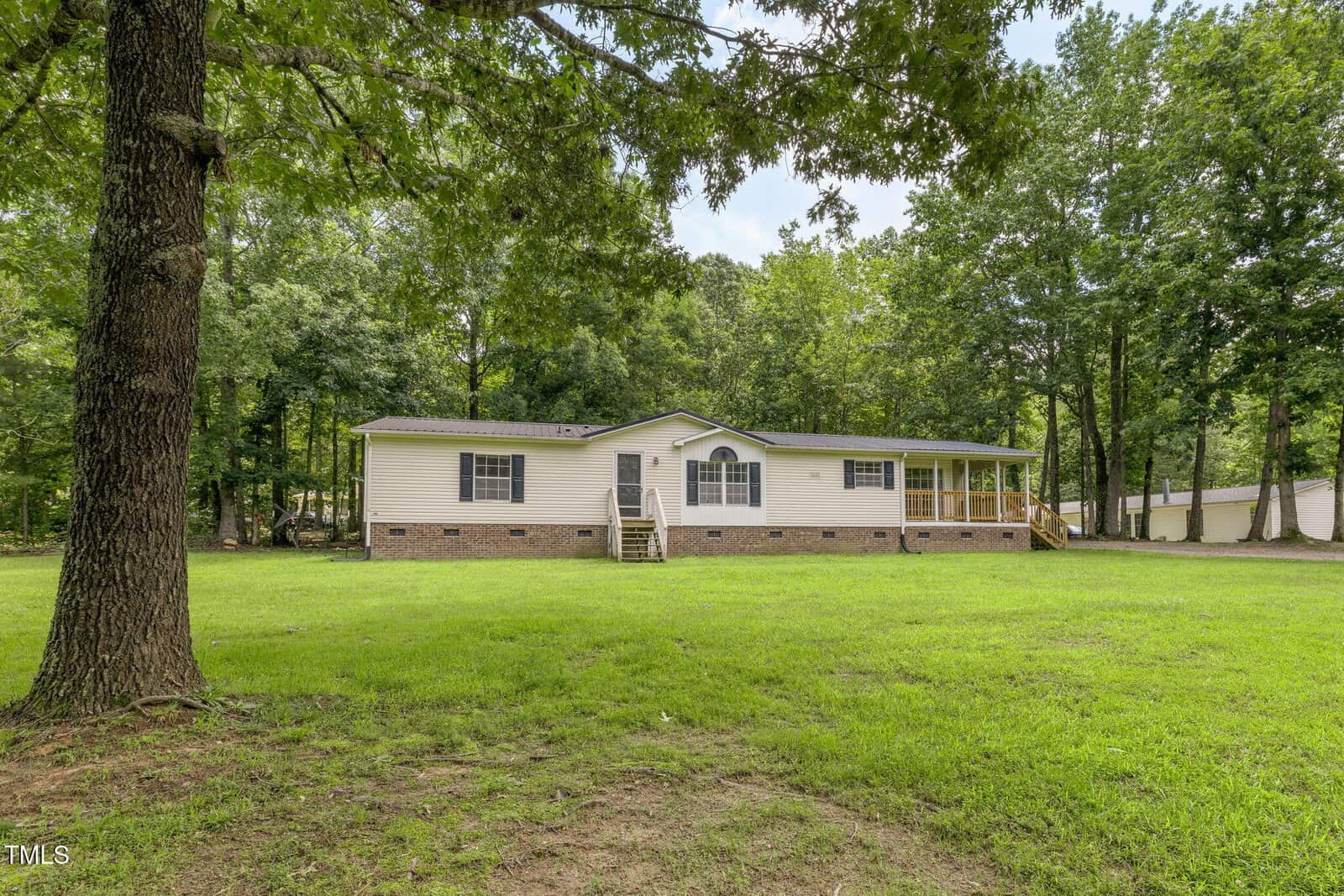 Single-story manufactured home with beige siding and a gable roof, set on a brick foundation. The house has a small front porch, steps leading to the front door, and is surrounded by green grass and trees.