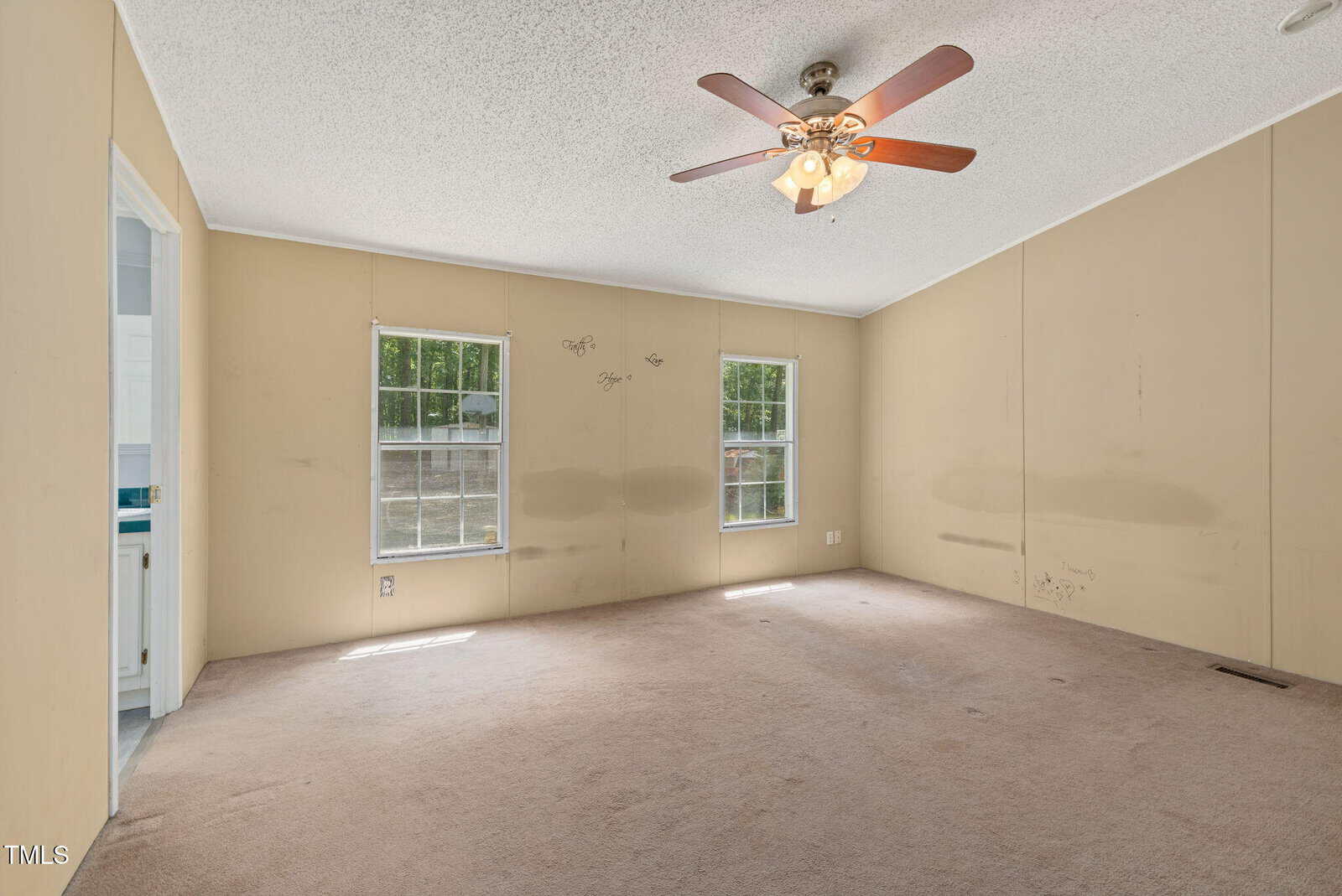 Empty beige-carpeted room with beige walls, two windows, a ceiling fan with lights, and an open doorway leading to a bathroom. Some wall marks and faint writing are visible.