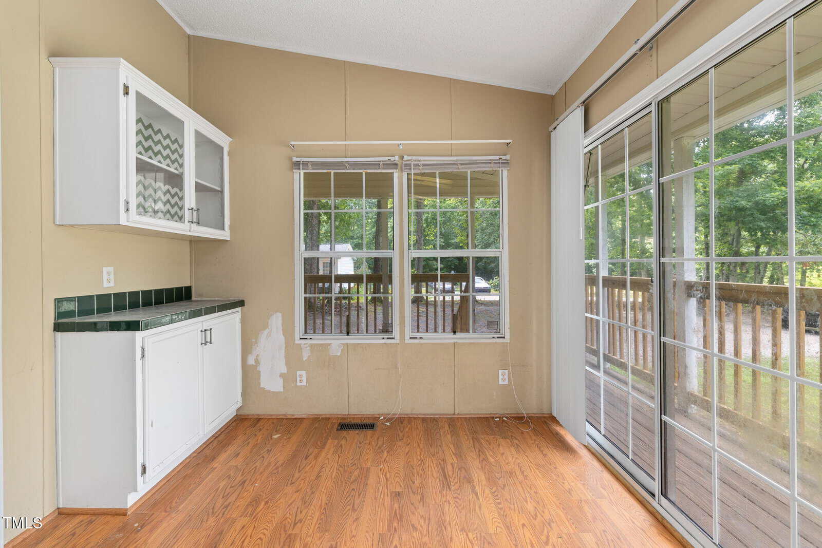 A small empty room with wood flooring, a built-in white cabinet, beige walls, two windows, and large sliding glass doors opening to a wooden deck with trees visible outside.