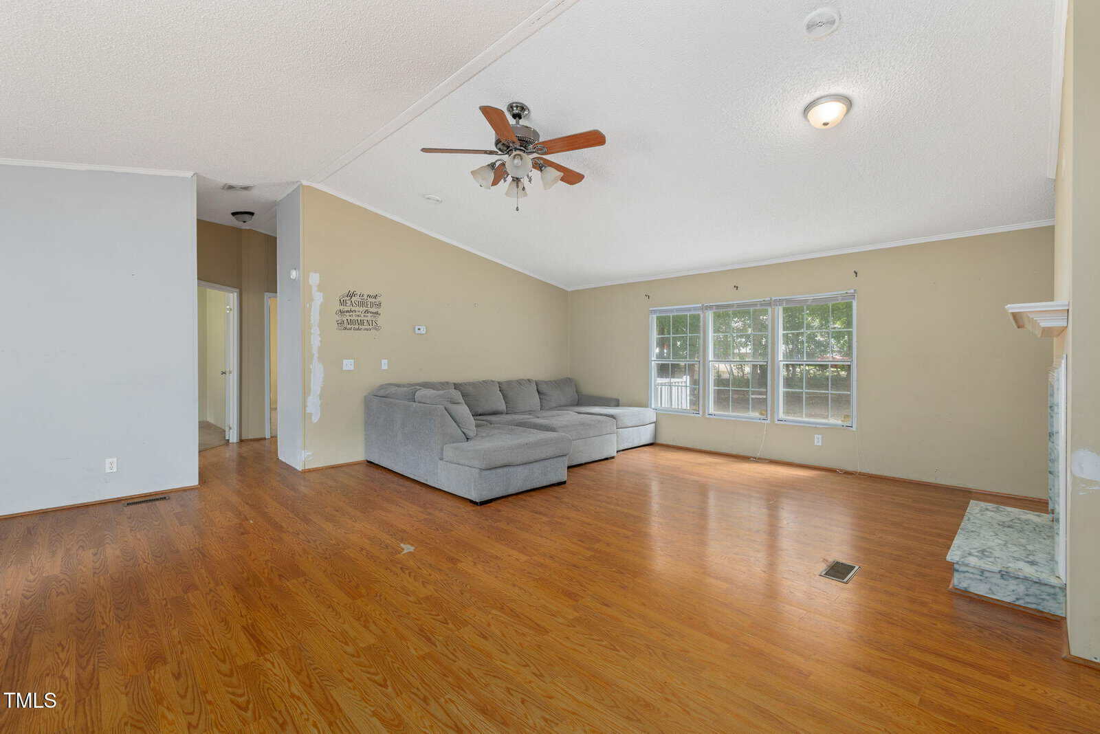 A spacious living room with light brown wood flooring, a gray sectional sofa, ceiling fan with lights, large windows letting in natural light, and beige walls with some visible paint patches.