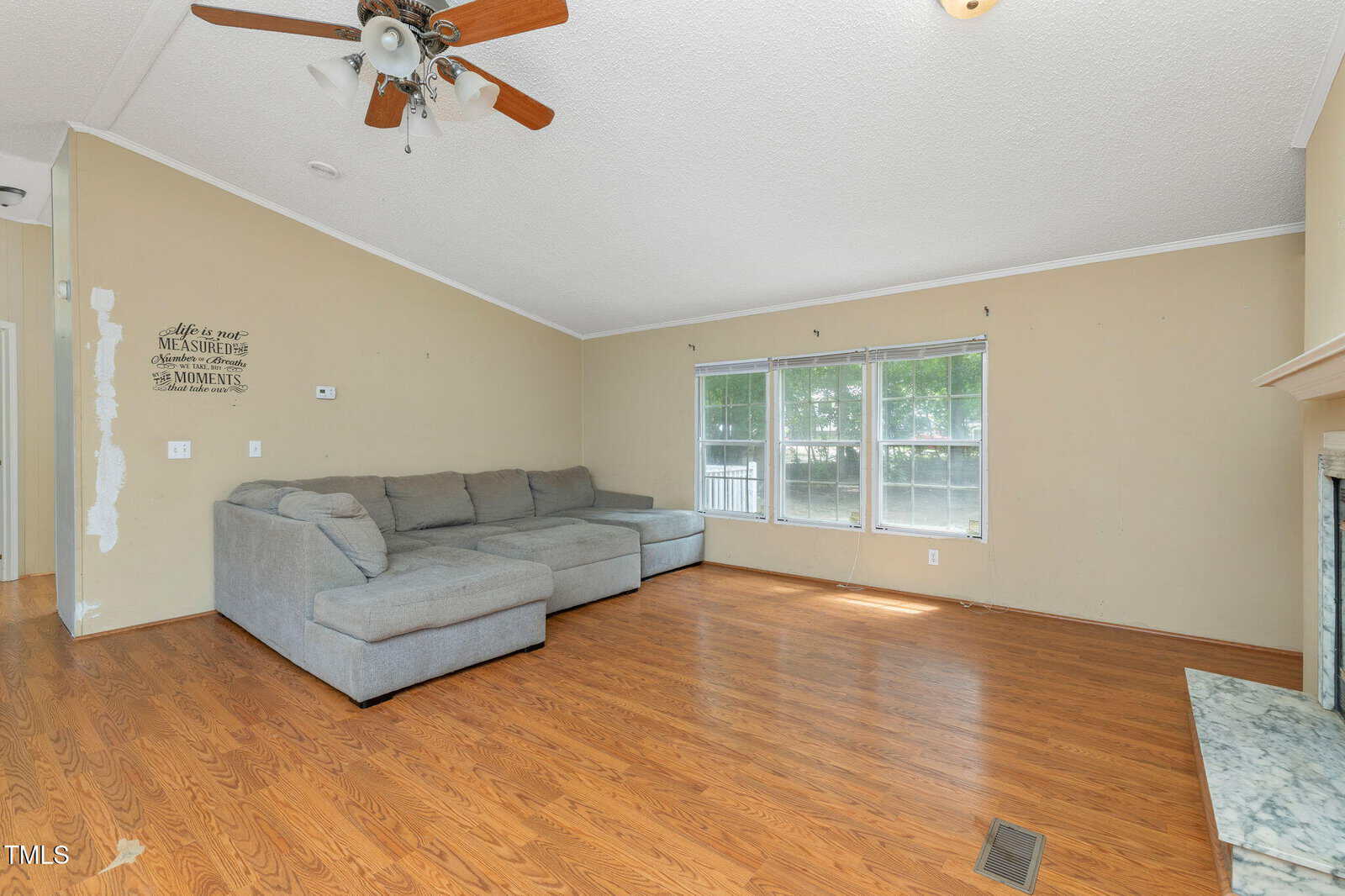 A living room with wood flooring, a large gray sectional sofa, three windows, a ceiling fan with lights, beige walls, and a fireplace on the right. Some wall paint is unfinished near the entrance.