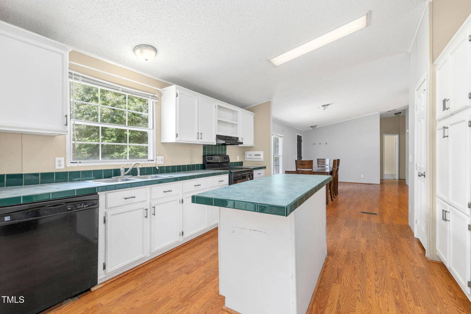 A kitchen with white cabinets, a green tiled countertop and island, black appliances, and wood flooring. A large window over the sink lets in natural light. The dining area is visible in the background.