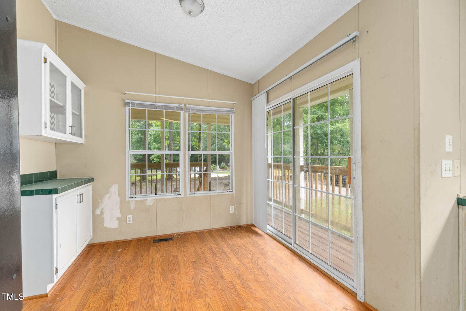 A small room with wood flooring, tan walls, large windows, and sliding glass doors leading to a covered deck. There are built-in white cabinets with green countertops on the left.