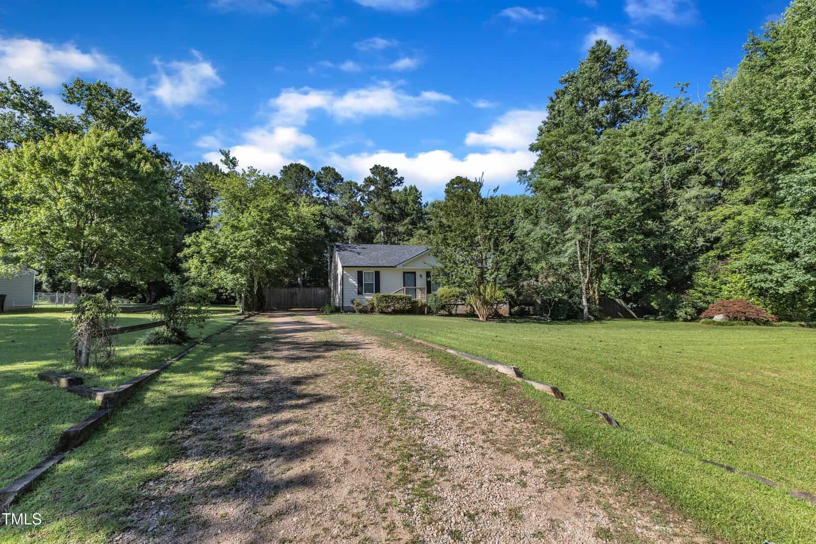 A small white house with a gray roof sits at the end of a long gravel driveway, surrounded by green grass, trees, and a clear blue sky.