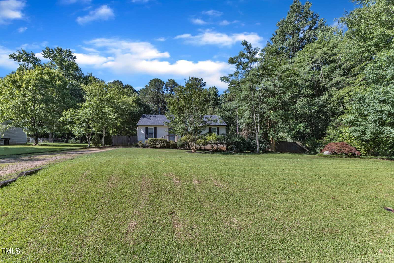 A small house surrounded by trees sits in the background, with a large, neatly mowed grassy lawn in front under a bright blue sky with some clouds.
