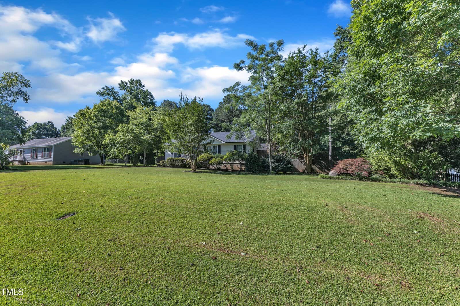 A spacious, well-maintained grassy yard with scattered mature trees and shrubs, featuring a single-story white house partially visible in the background under a blue sky with clouds.