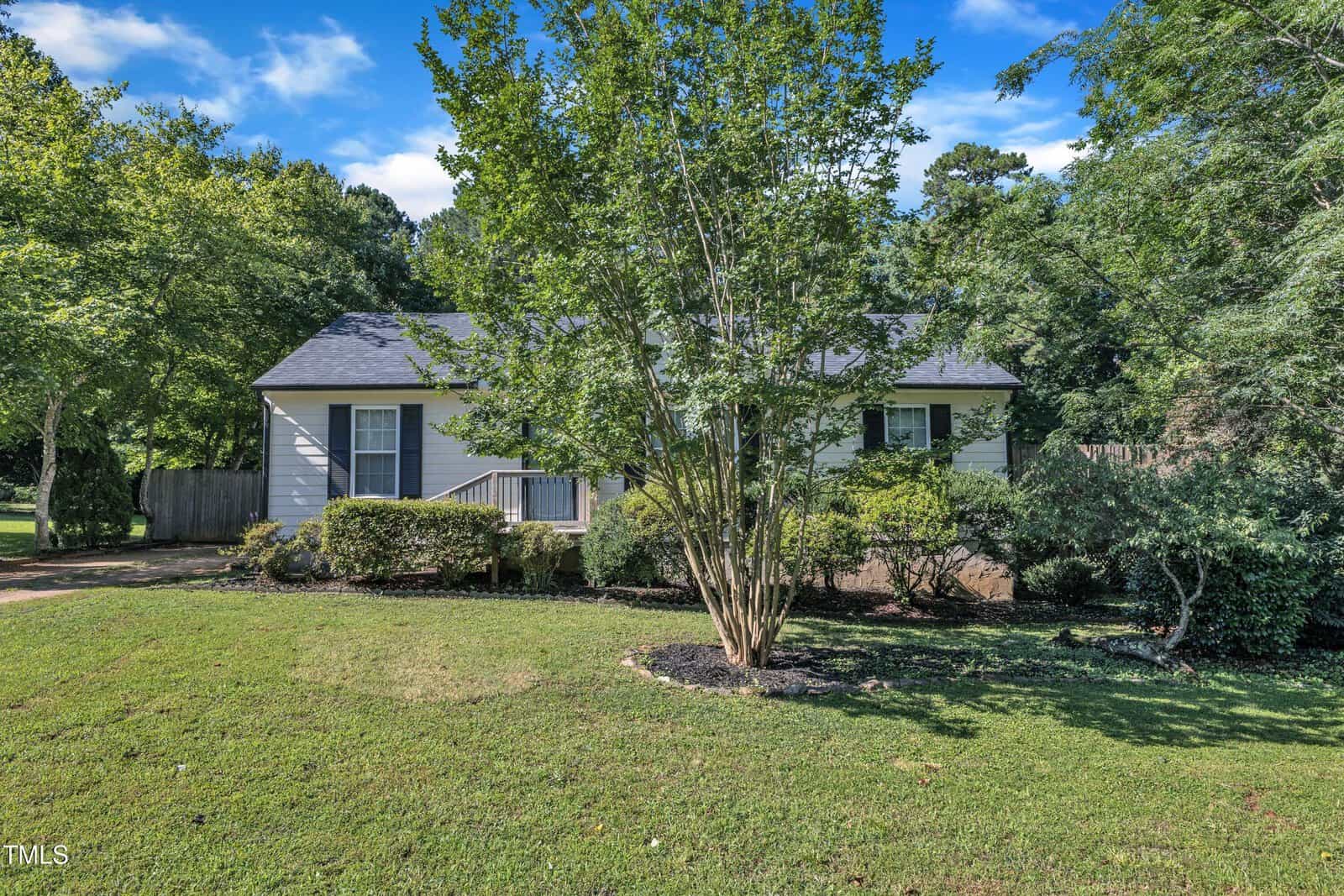 Single-story white house with dark roof partially hidden behind a leafy tree, surrounded by green grass, shrubs, and trees under a blue sky with scattered clouds.