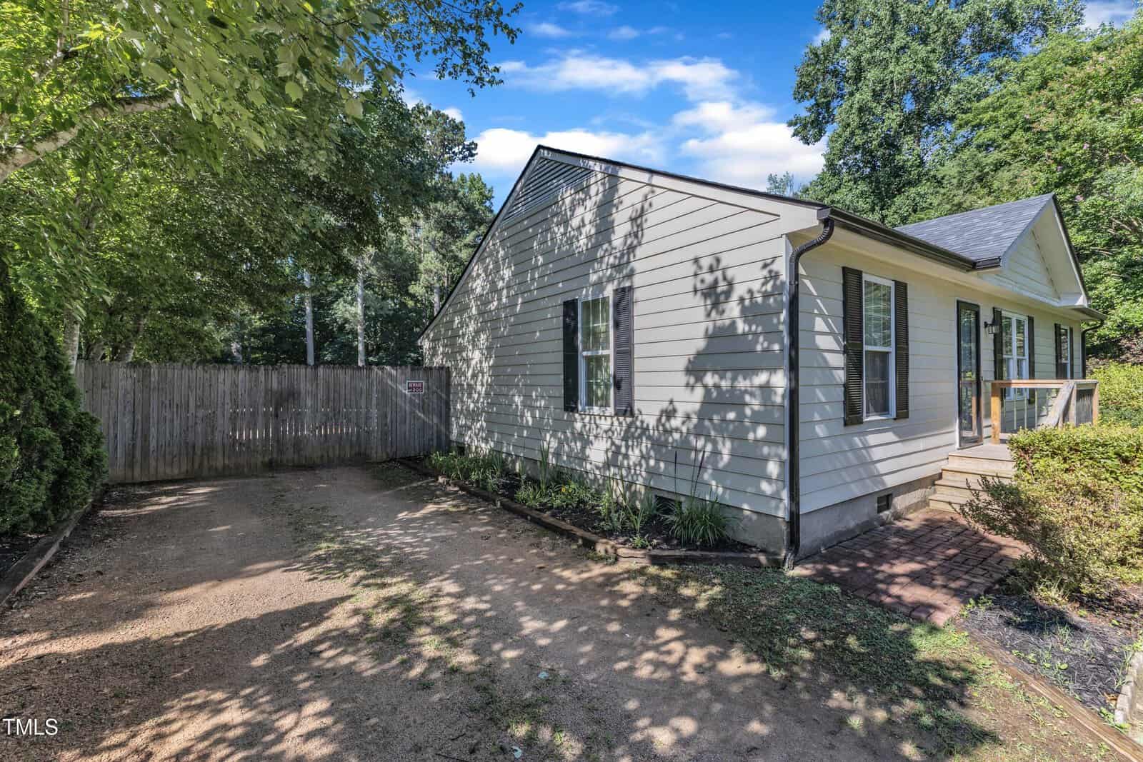 Single-story house with light-colored siding, black shutters, and a shingled roof. There is a gravel driveway, wooden fence, brick walkway, and green trees and shrubs around the house under a blue sky with clouds.