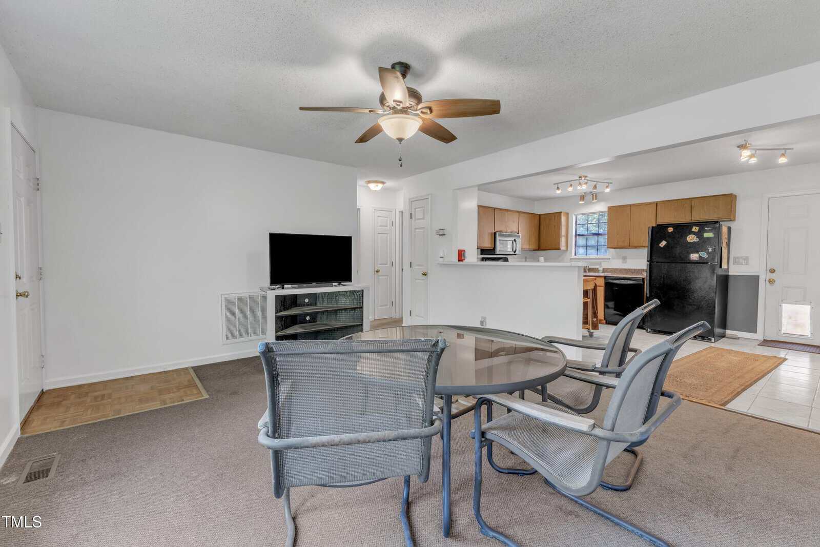 A living area with a ceiling fan, glass table with four metal chairs, TV on a stand, and an open kitchen featuring wooden cabinets, black appliances, and a countertop with stools. The walls are white and the floor is carpeted.