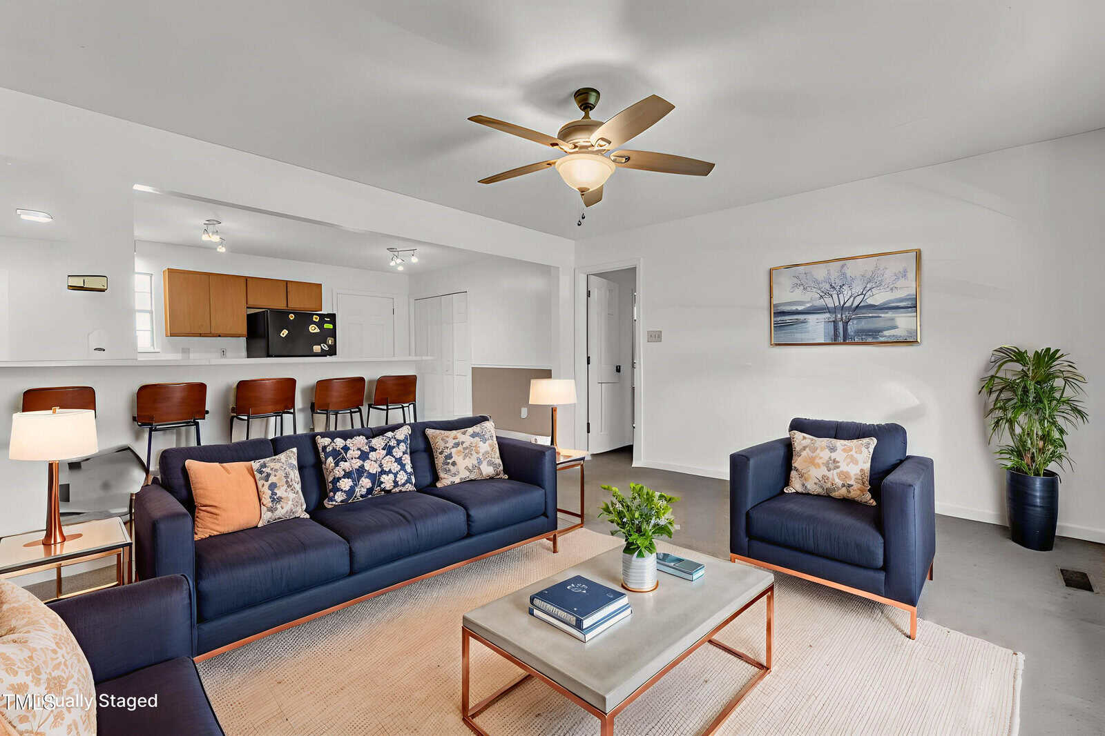 A modern living room with two navy sofas, a beige armchair, a coffee table with books and a plant, a ceiling fan, and artwork on the wall. The kitchen and dining area are visible in the background.