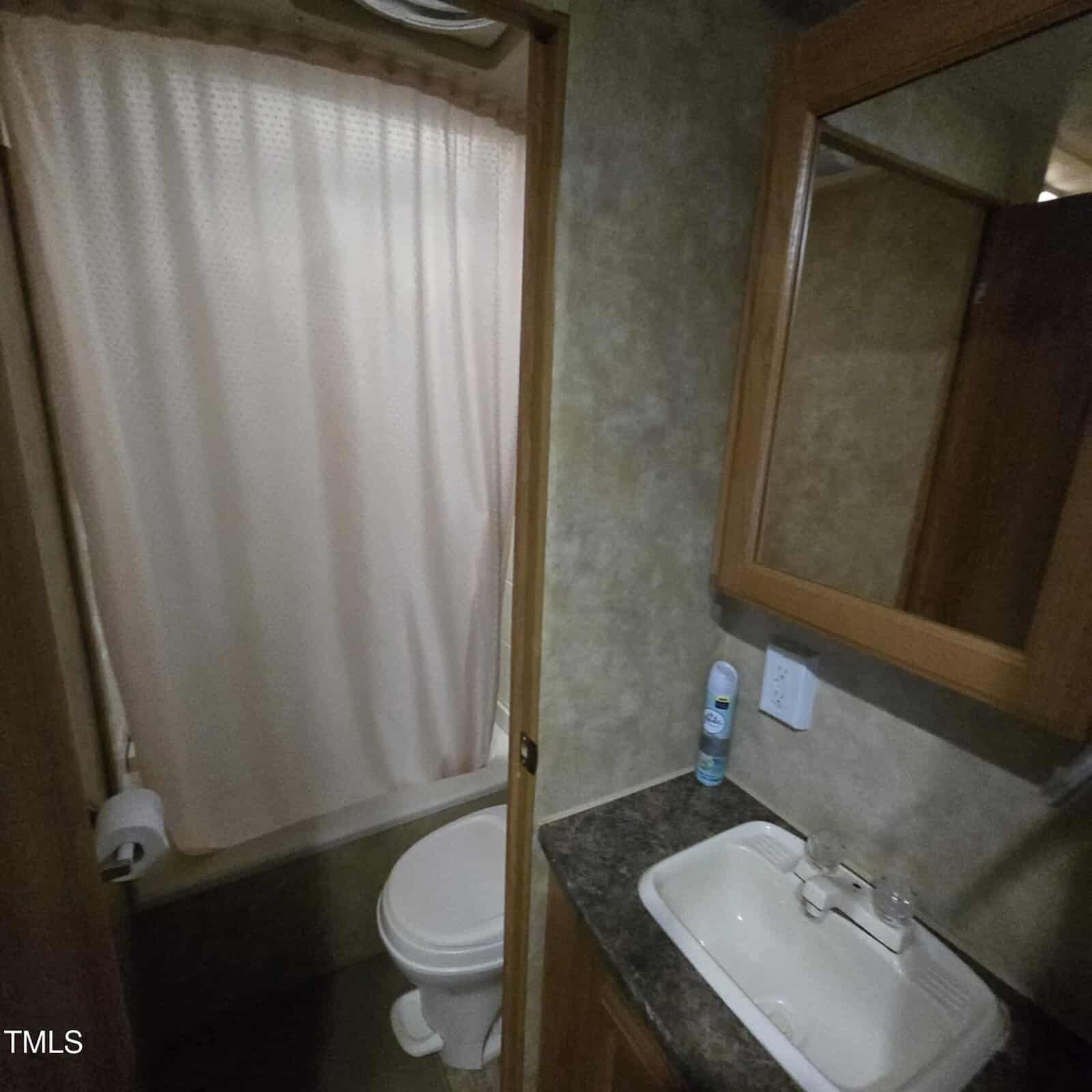 Small bathroom with a shower and curtain, a toilet, a sink with a countertop, a wall-mounted mirror cabinet, a soap dispenser, and a can of air freshener on the counter. Walls have a beige, textured appearance.