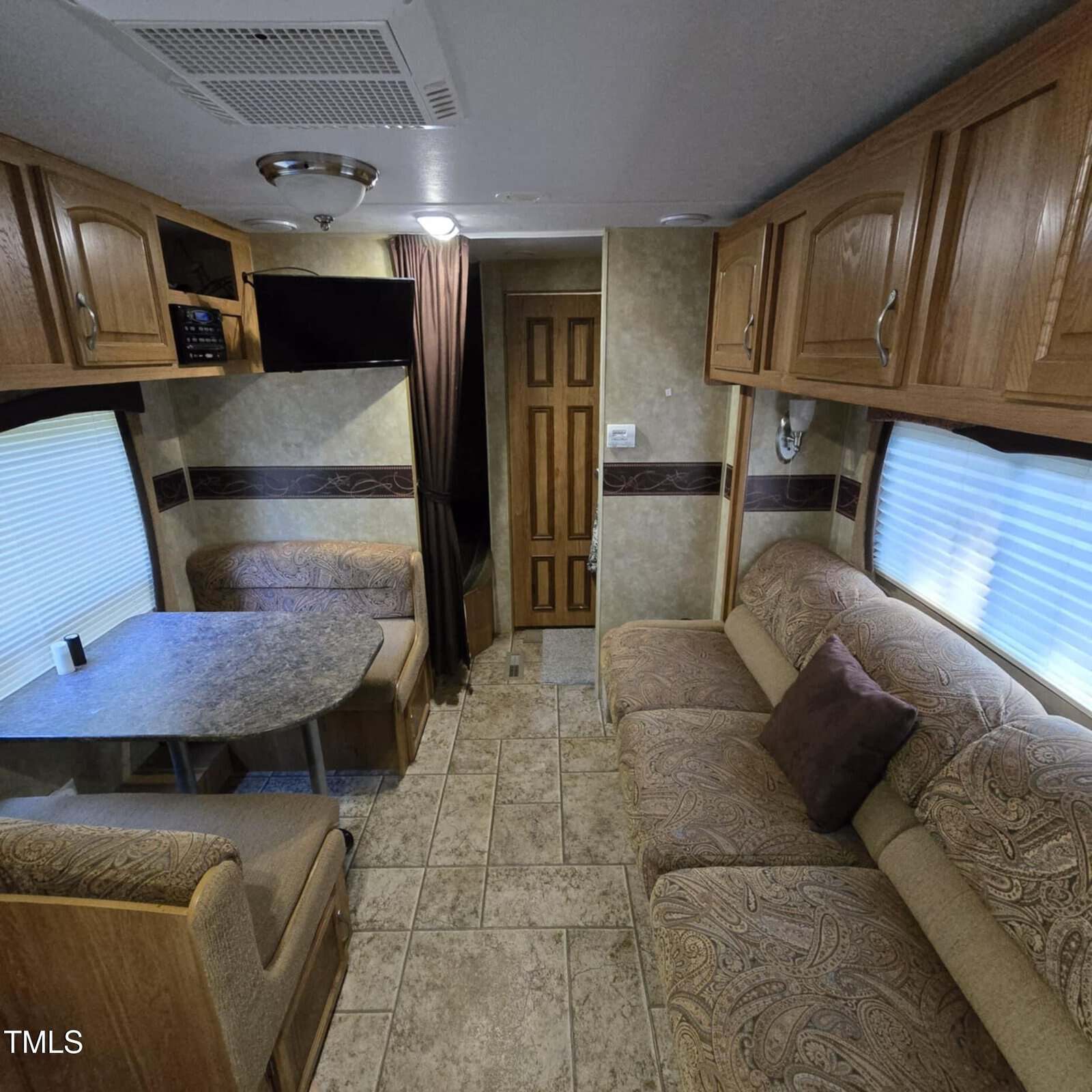 Interior of an RV showing a dining area with a table and benches on the left, a couch with a pillow on the right, wood cabinets above, tiled flooring, and a door at the back. The walls are beige with brown trim.