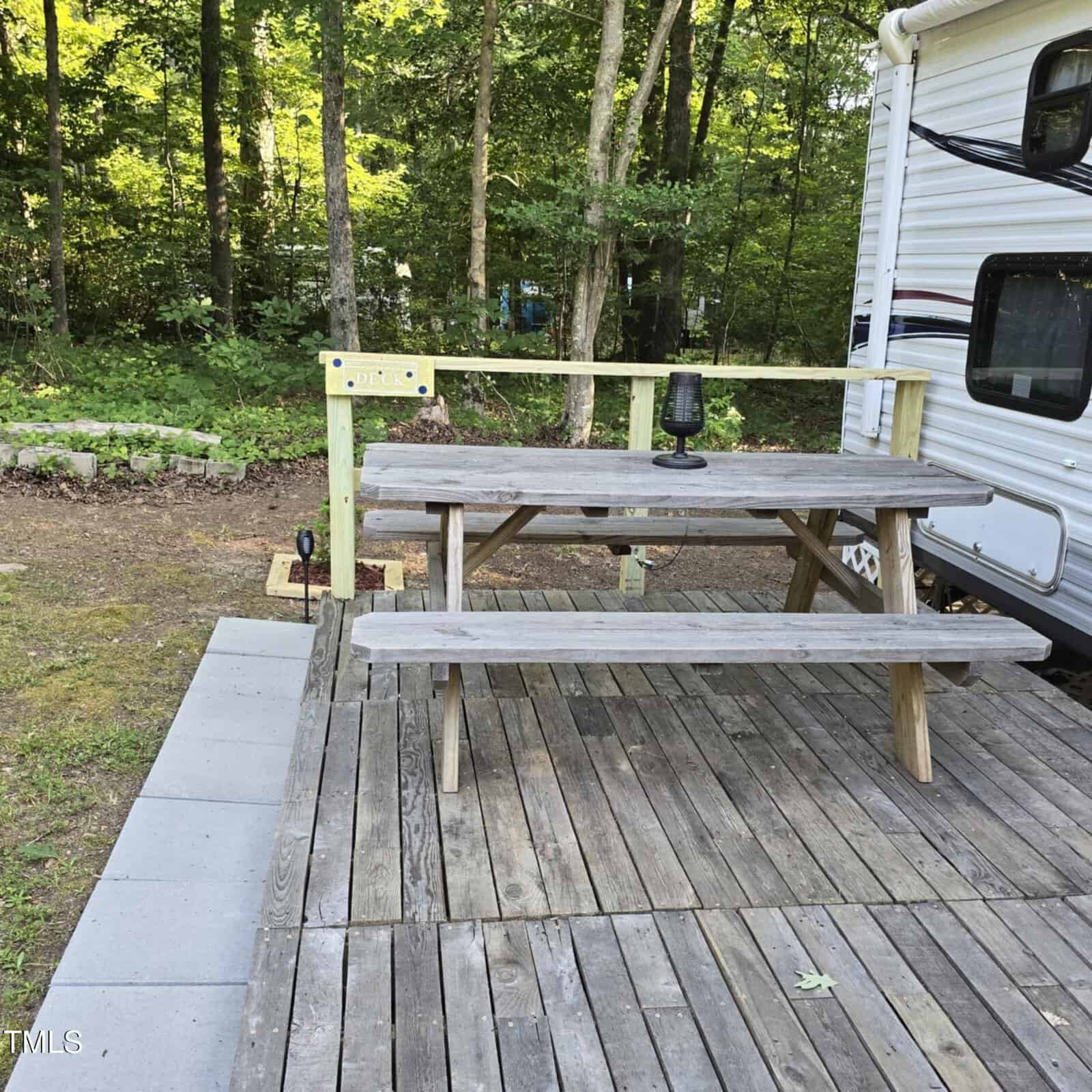 A wooden picnic table sits on a wooden deck next to a white camper in a wooded area, with a small lamp on the table and stone pavers leading to the deck.