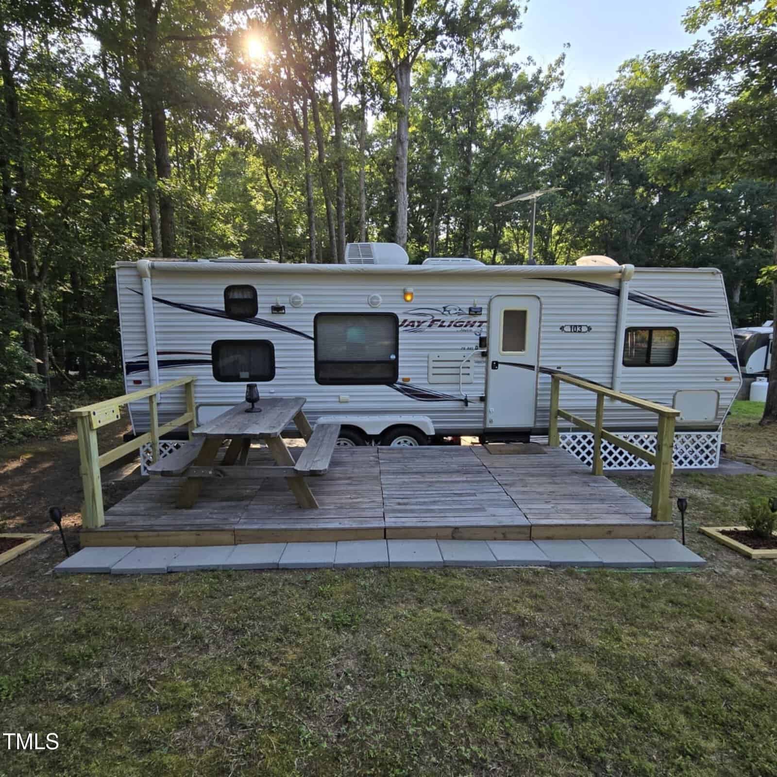A white travel trailer is parked in a wooded area. In front, there is a wooden deck with a picnic table and simple railings. Sunlight filters through the trees in the background.
