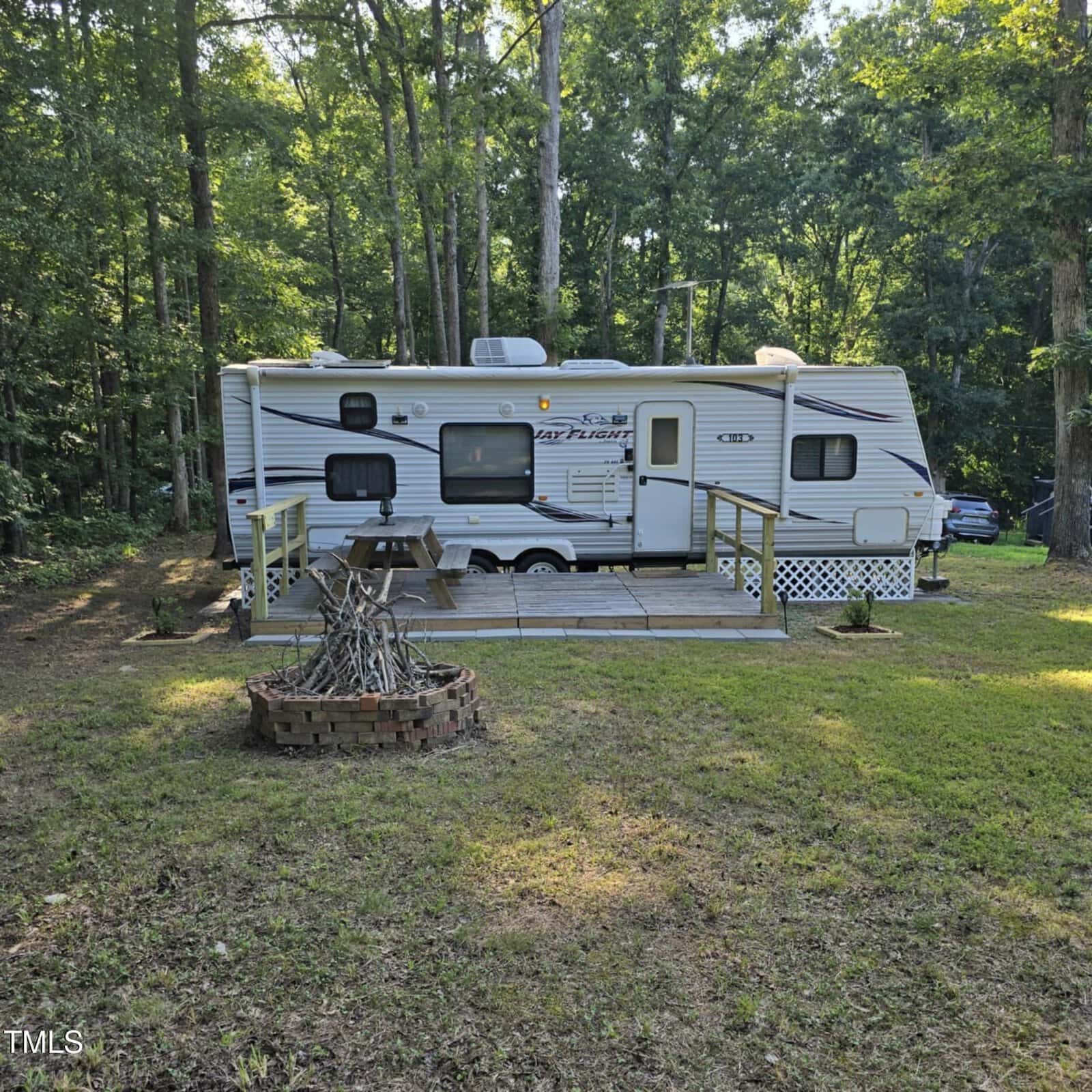 A white travel trailer with a small wooden deck sits in a wooded area surrounded by trees. There is a fire pit made of bricks in the grassy yard in front of the trailer, and sunlight filters through the leaves.