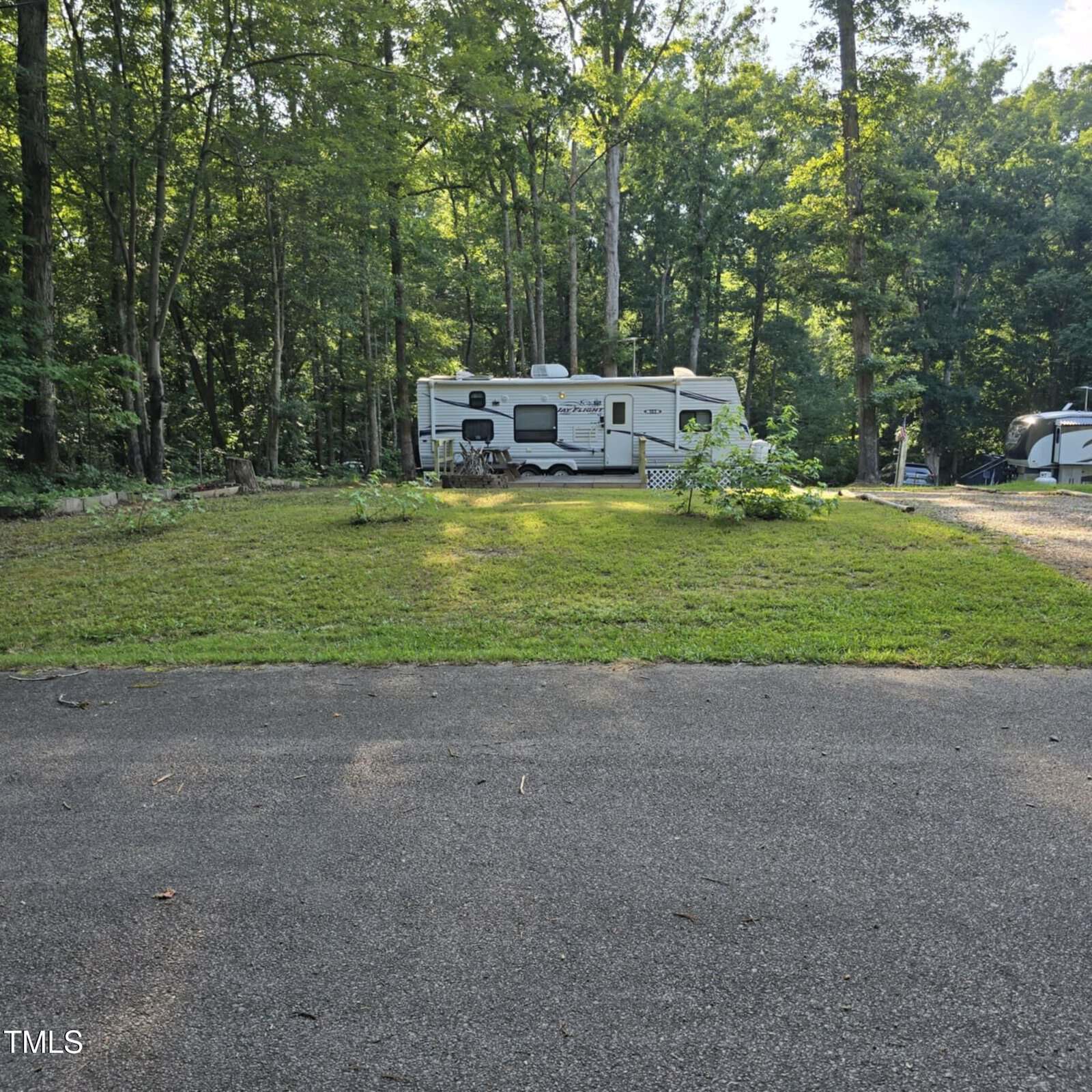 A white RV is parked on a grassy lot surrounded by tall trees in a wooded area, with a paved area in the foreground and another trailer partially visible to the right.