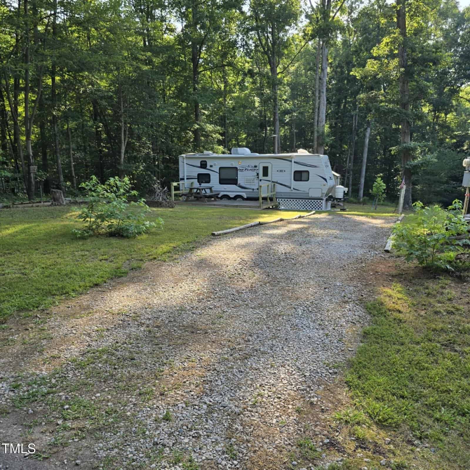 A gravel driveway leads to a white travel trailer parked in a clearing surrounded by tall trees and green grass on a sunny day.