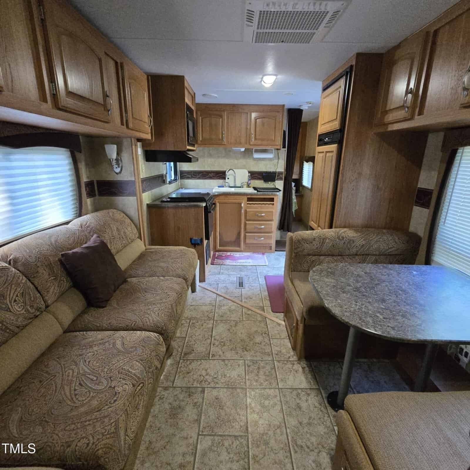 Interior of an RV showing a patterned sofa, a small dining table with two seats, kitchen with stove and sink, wooden cabinets, tiled floor, and a window with blinds on each side. Lighting is soft and natural.