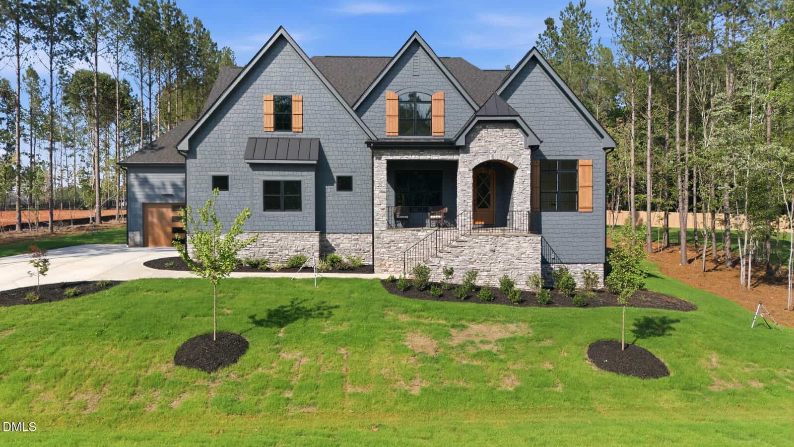 A modern gray house with stone accents, wood shutters, and a front porch, set on a green lawn with young trees and surrounded by tall pine trees under a blue sky.