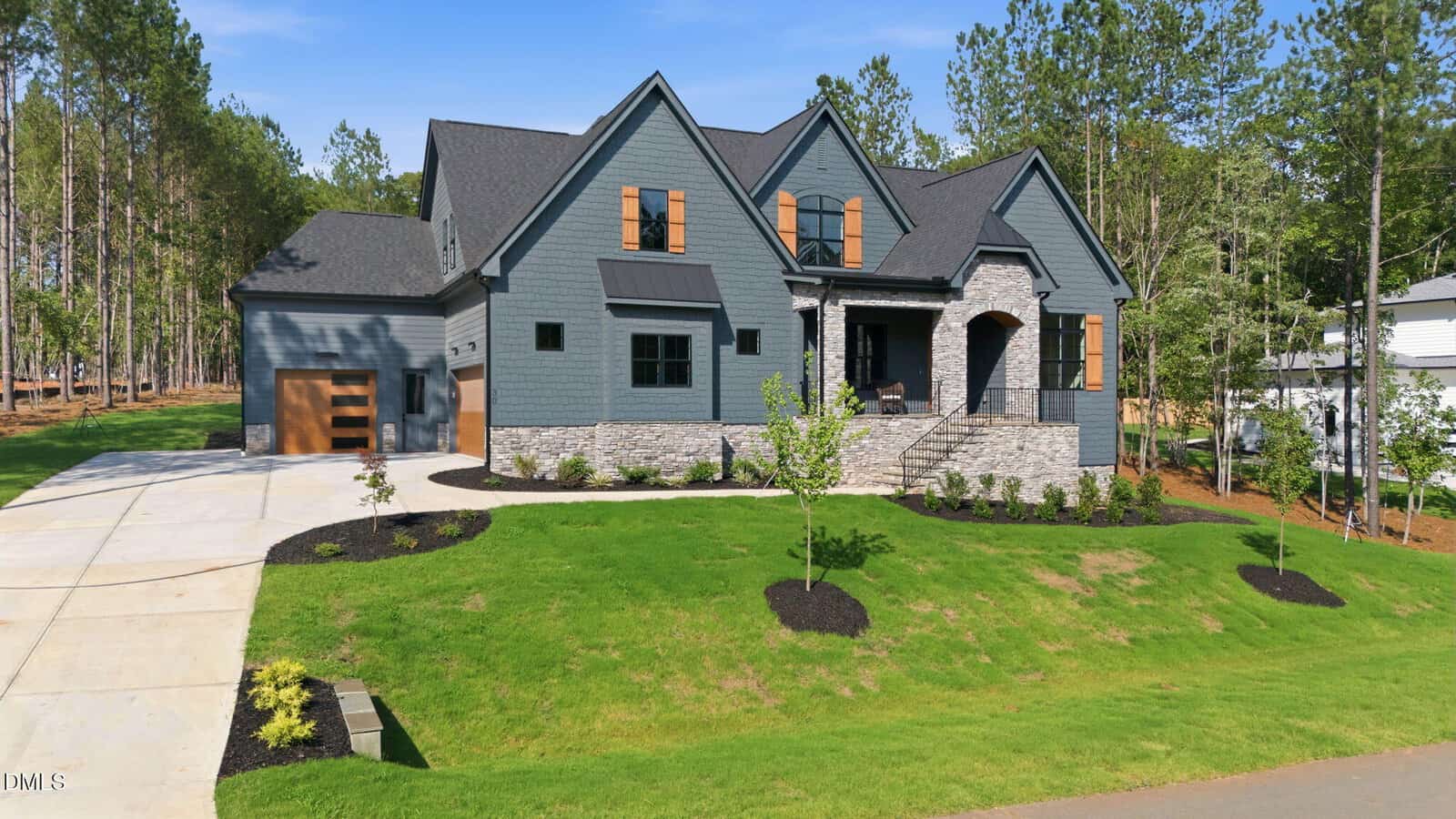 Modern two-story house with blue-gray siding, stone accents, and a black roof. It features a covered porch, steps leading to the entrance, a two-car garage, and a neatly landscaped green yard with young trees.
