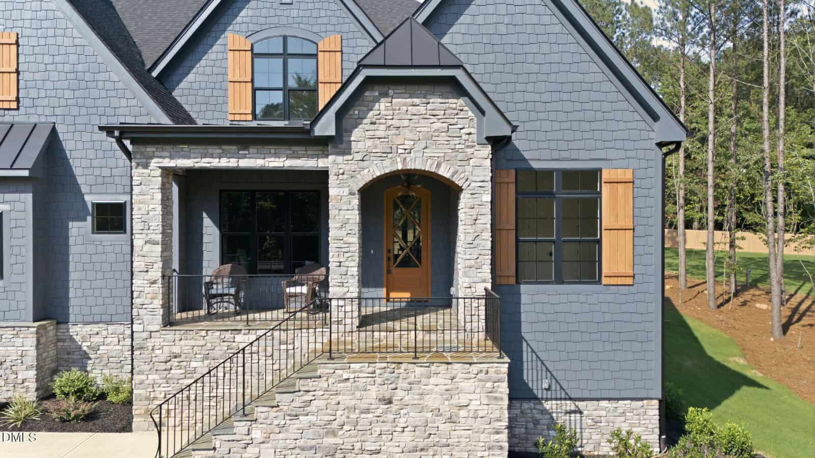Gray stone house with wooden shutters, a wooden front door, a small porch with two chairs, black railings, steps leading up, and green grass with trees in the background.