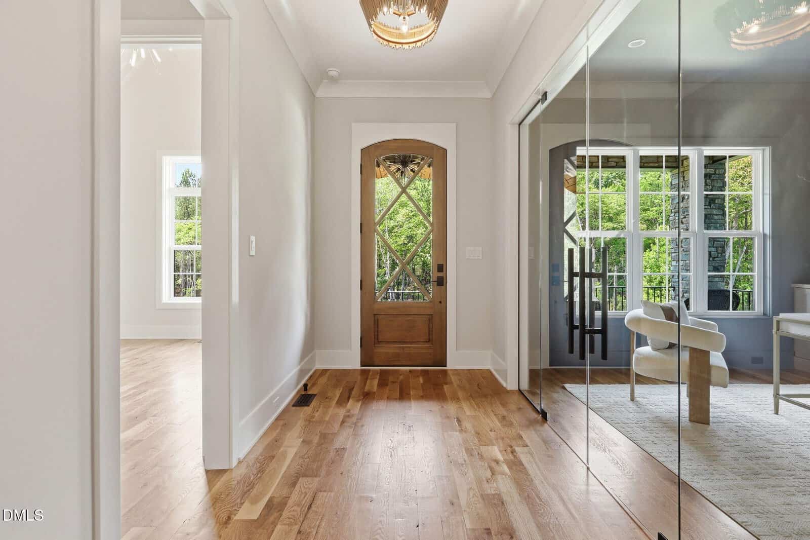A bright hallway with wooden floors, a wooden front door with glass panels, white walls, and a glass-walled room with a modern chair and desk on the right. Large windows let in natural light and show green trees outside.