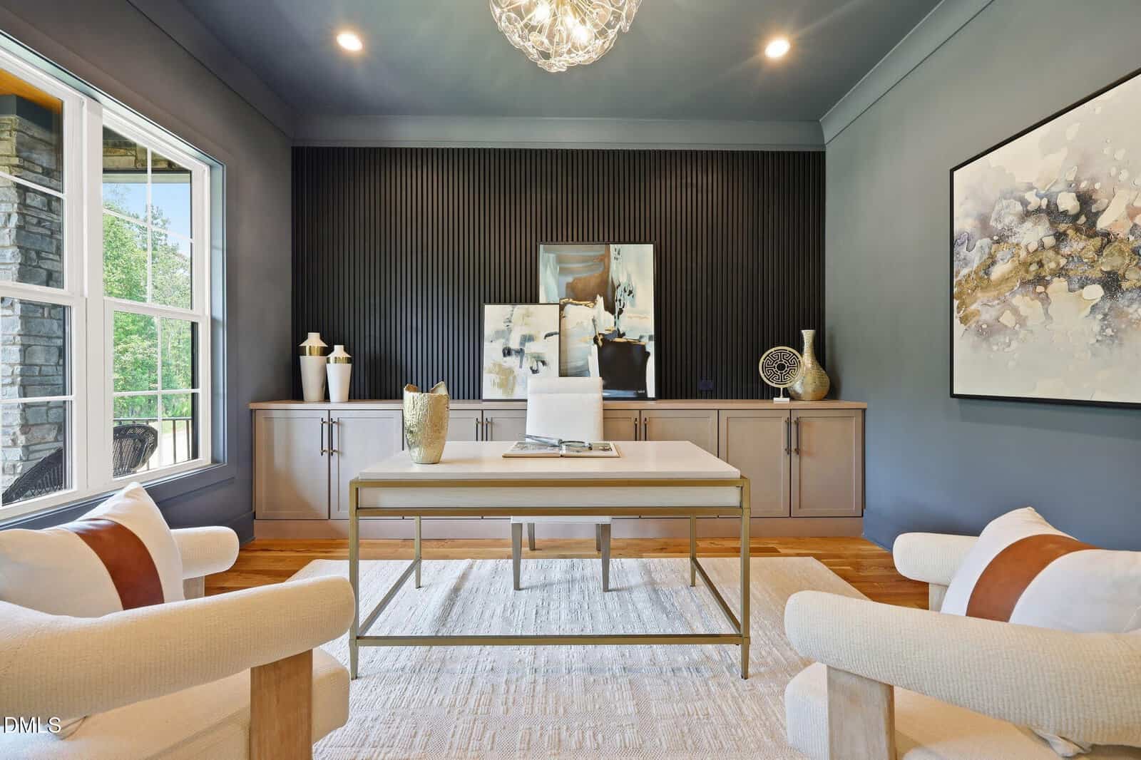 Modern home office with a glass-top desk, white chair, two beige armchairs, abstract wall art, and decor on a sideboard. Dark accent wall and large window provide contrast and natural light.
