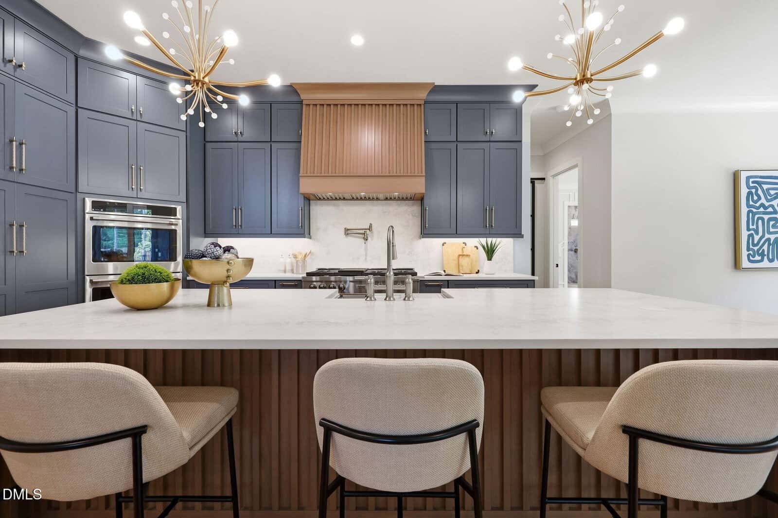 Modern kitchen with a large marble island, three beige chairs, blue-gray cabinets, gold sconces, stainless steel appliances, and decorative items on the counter. Wall art is visible on the right side.