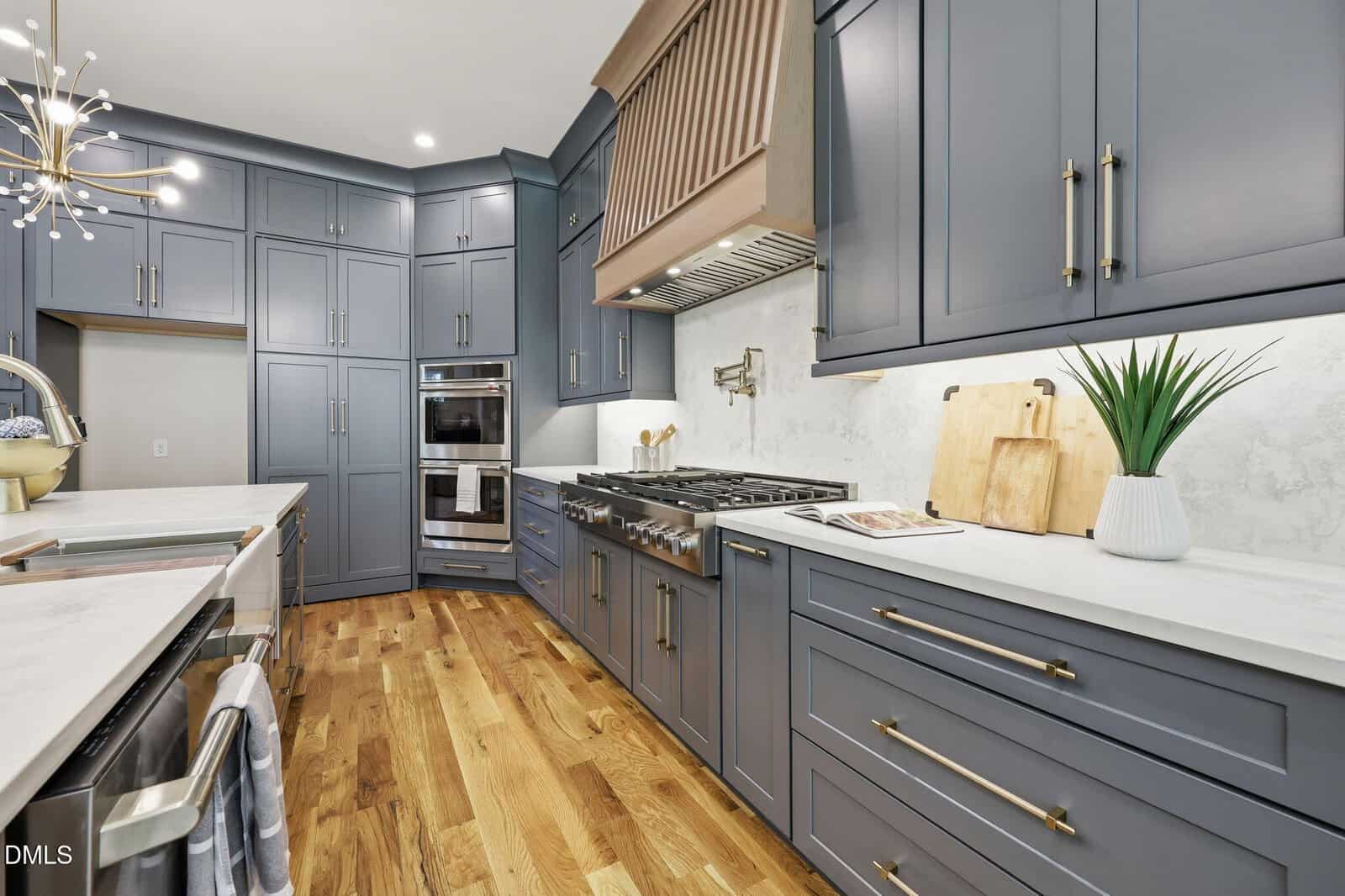 Modern kitchen with blue-gray cabinets, white countertops, stainless steel appliances, wood flooring, and gold hardware. A stove with a large hood is in the center. A plant and cutting boards are on the right counter.