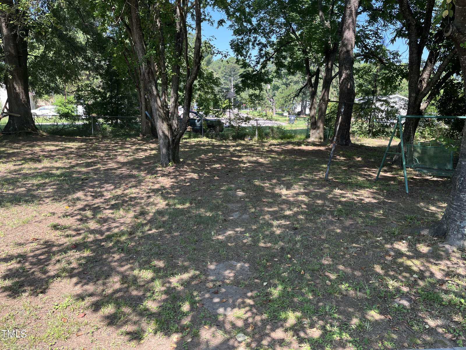 A backyard with patchy grass and dirt, scattered shade from several large trees, a swing set frame on the right, and some cars and houses visible in the background.