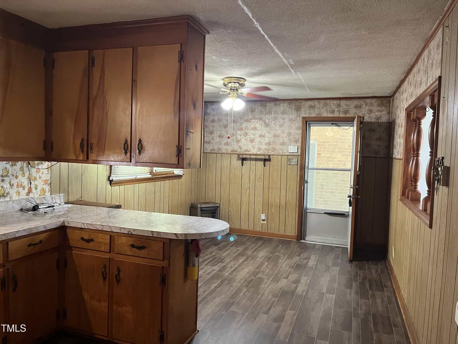 A kitchen with wooden cabinets, marble-pattern countertops, and wood-paneled walls leads into a dining area with a ceiling fan, door to the outside, and floral wallpaper. The floor is dark wood laminate.