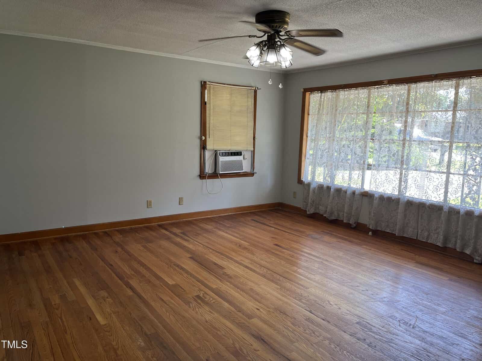 Empty room with hardwood floors, light gray walls, a ceiling fan with lights, a window with an air conditioning unit, and a large window with sheer curtains letting in natural light.