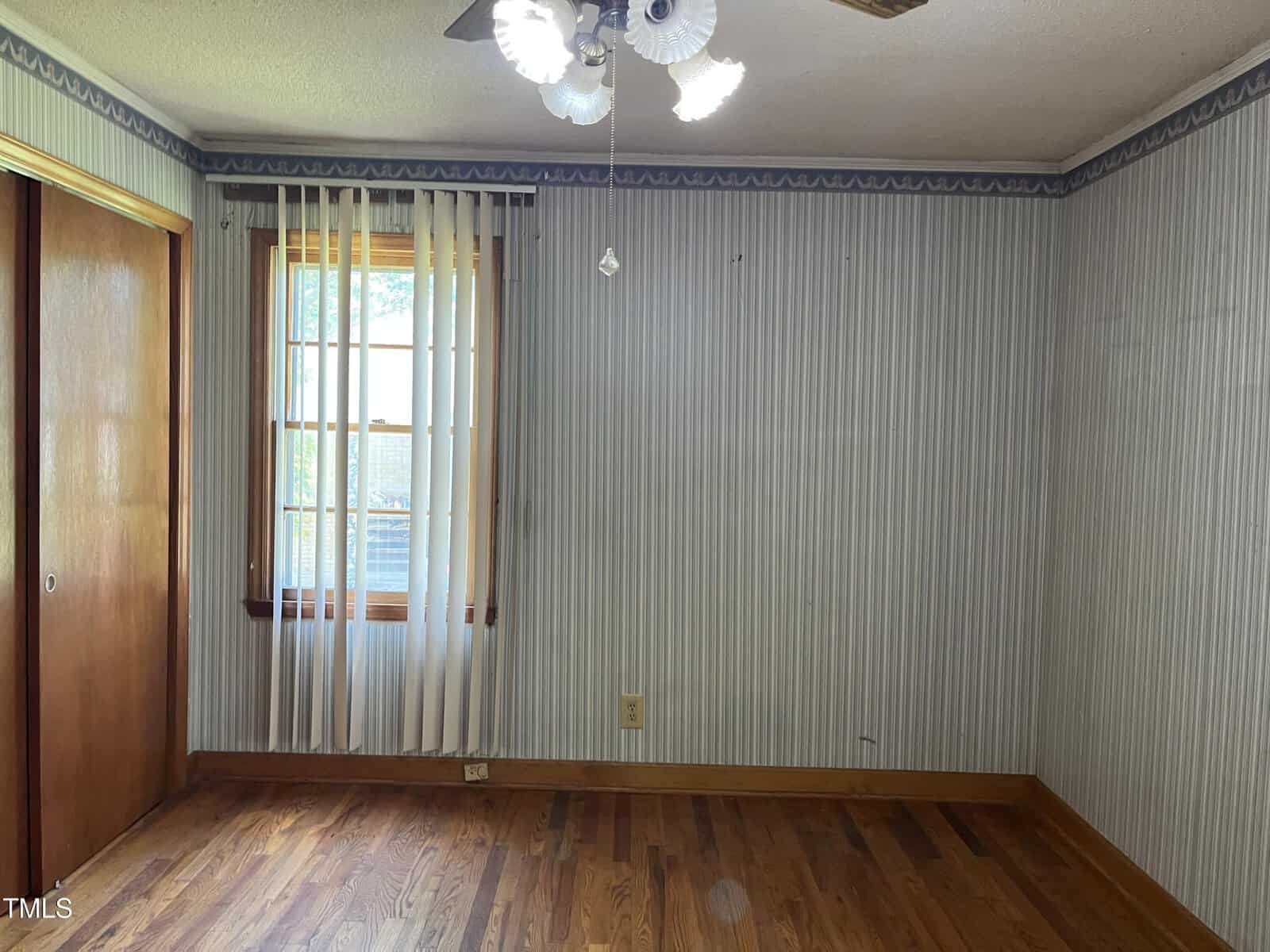 Empty room with hardwood floor, striped wallpaper, a window with vertical blinds, wooden closet doors, and a ceiling fan with four lights. Sunlight enters through the window, illuminating the space.