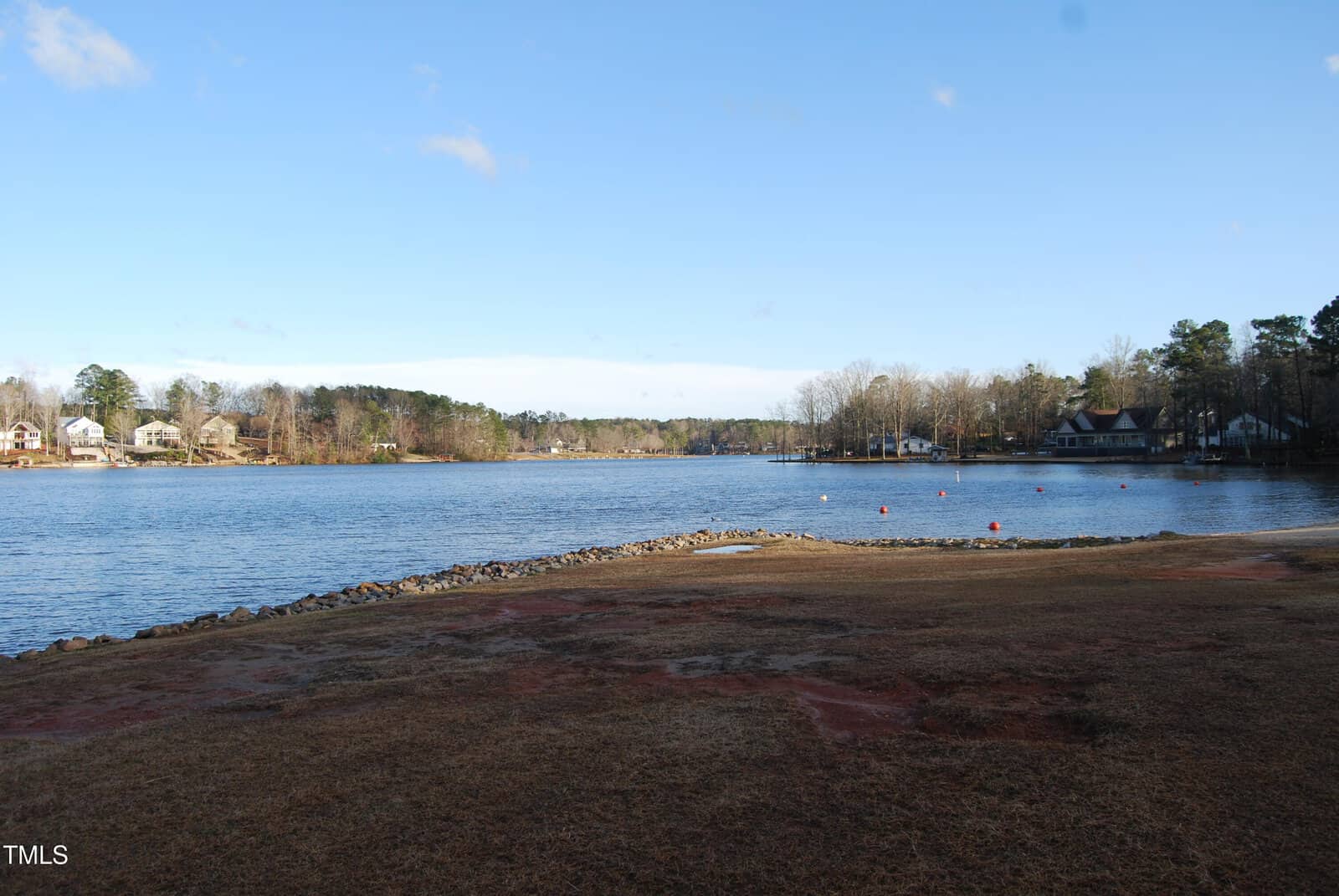 A calm lake bordered by grass and rocks, with houses and leafless trees lining the far shore under a clear blue sky.