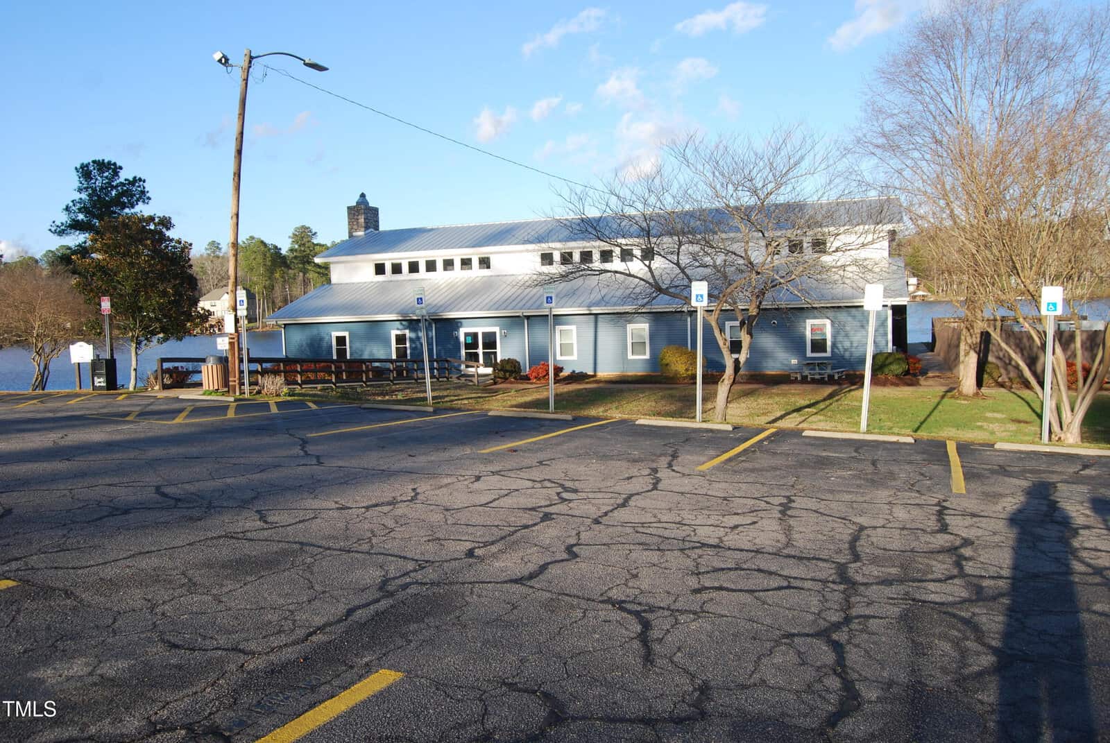 A one-story light blue building with a metal roof sits behind an empty, cracked parking lot with marked accessible spaces. Leafless trees line the front; a lake and trees are visible in the background under a clear sky.
