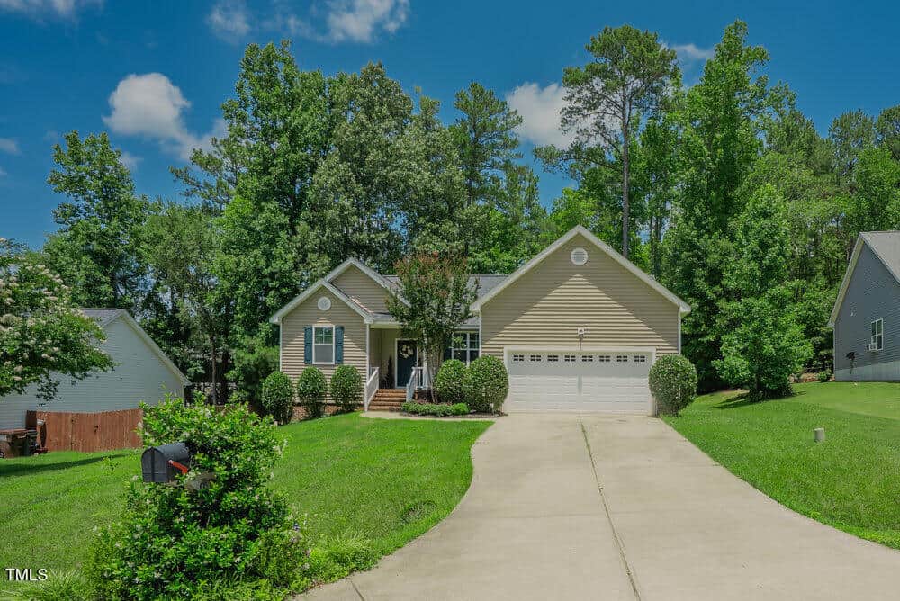 Single-story beige house with a two-car garage, front porch, and well-maintained yard surrounded by green grass, trees, and shrubs, set on a sunny day with a blue sky and a few clouds.