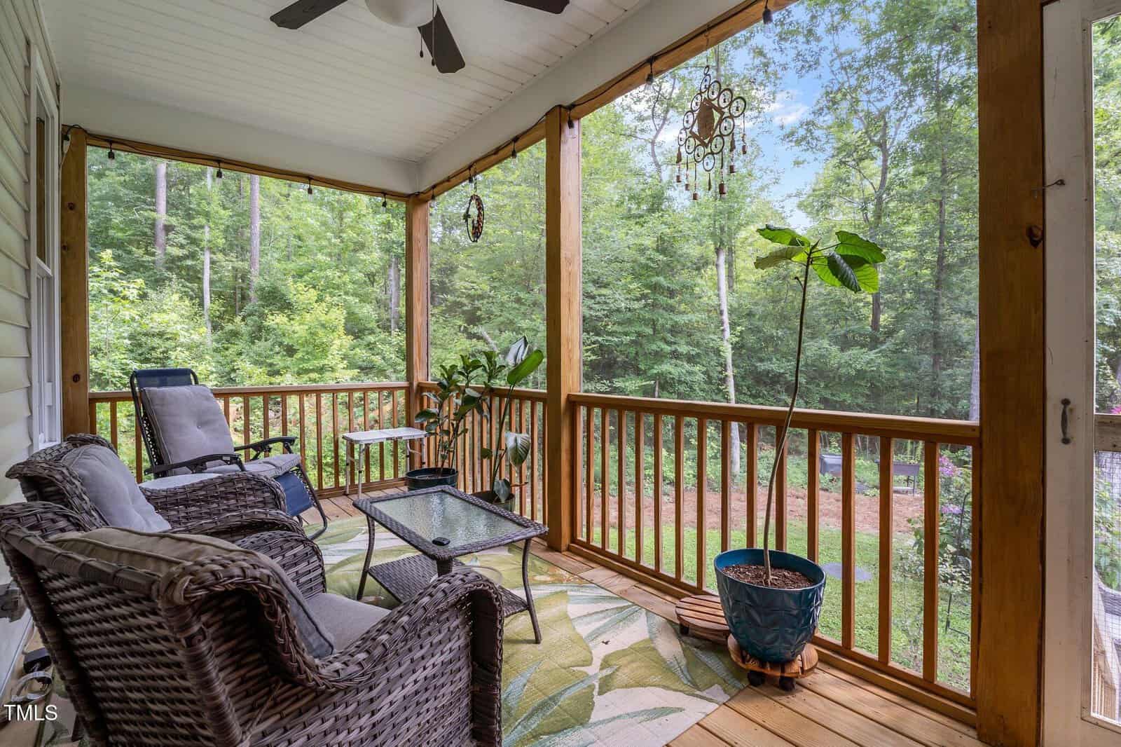 A cozy screened-in porch with wicker chairs, a glass-top table, potted plants, and a leafy rug overlooks a green, wooded yard. The space has wood railings and hanging decorations.