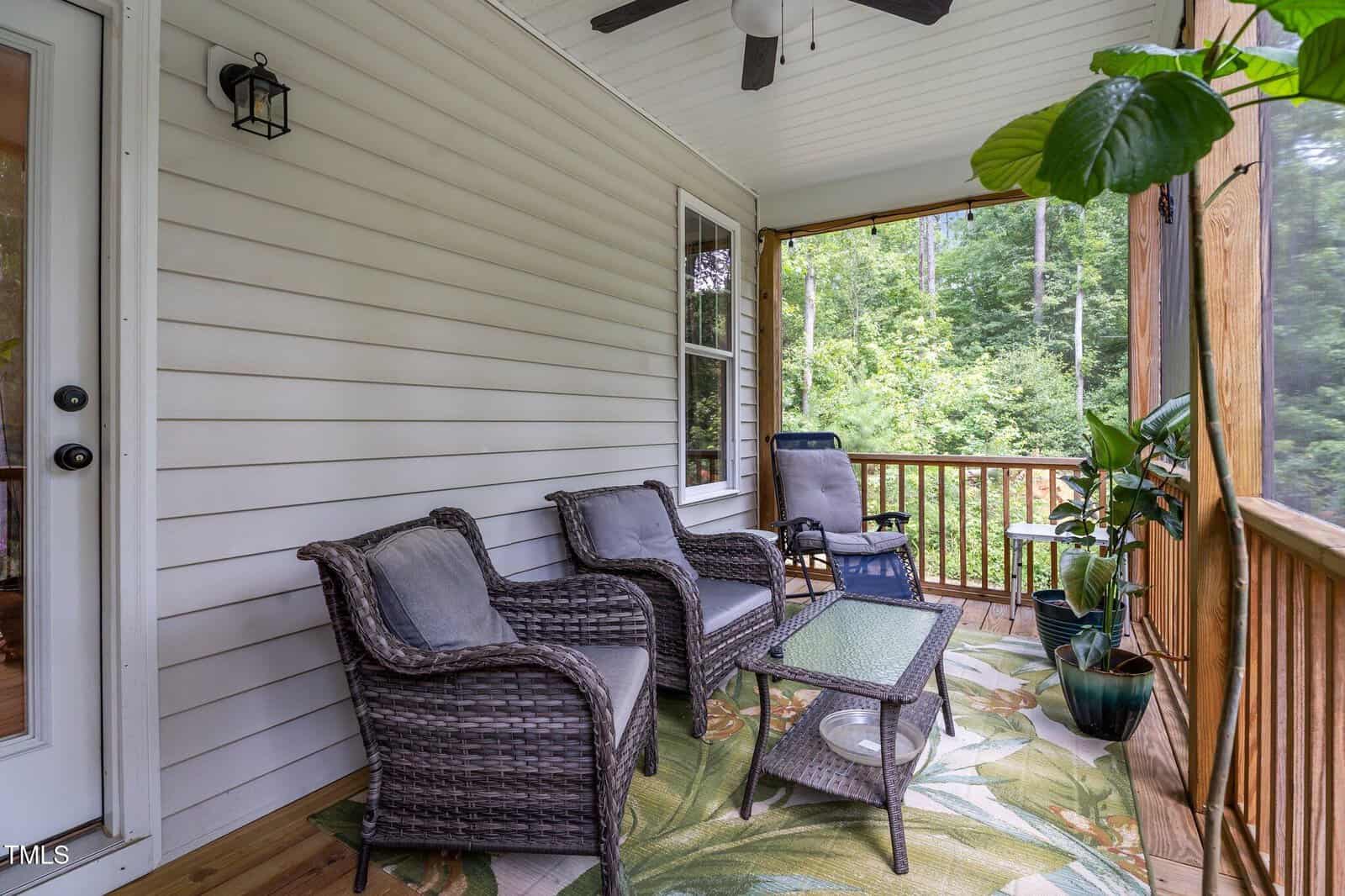 A screened-in porch with wicker chairs and a glass-top table, a lounge chair, potted plants, a ceiling fan, and a leafy outdoor rug. The porch overlooks green trees and is enclosed with wood railings and screens.