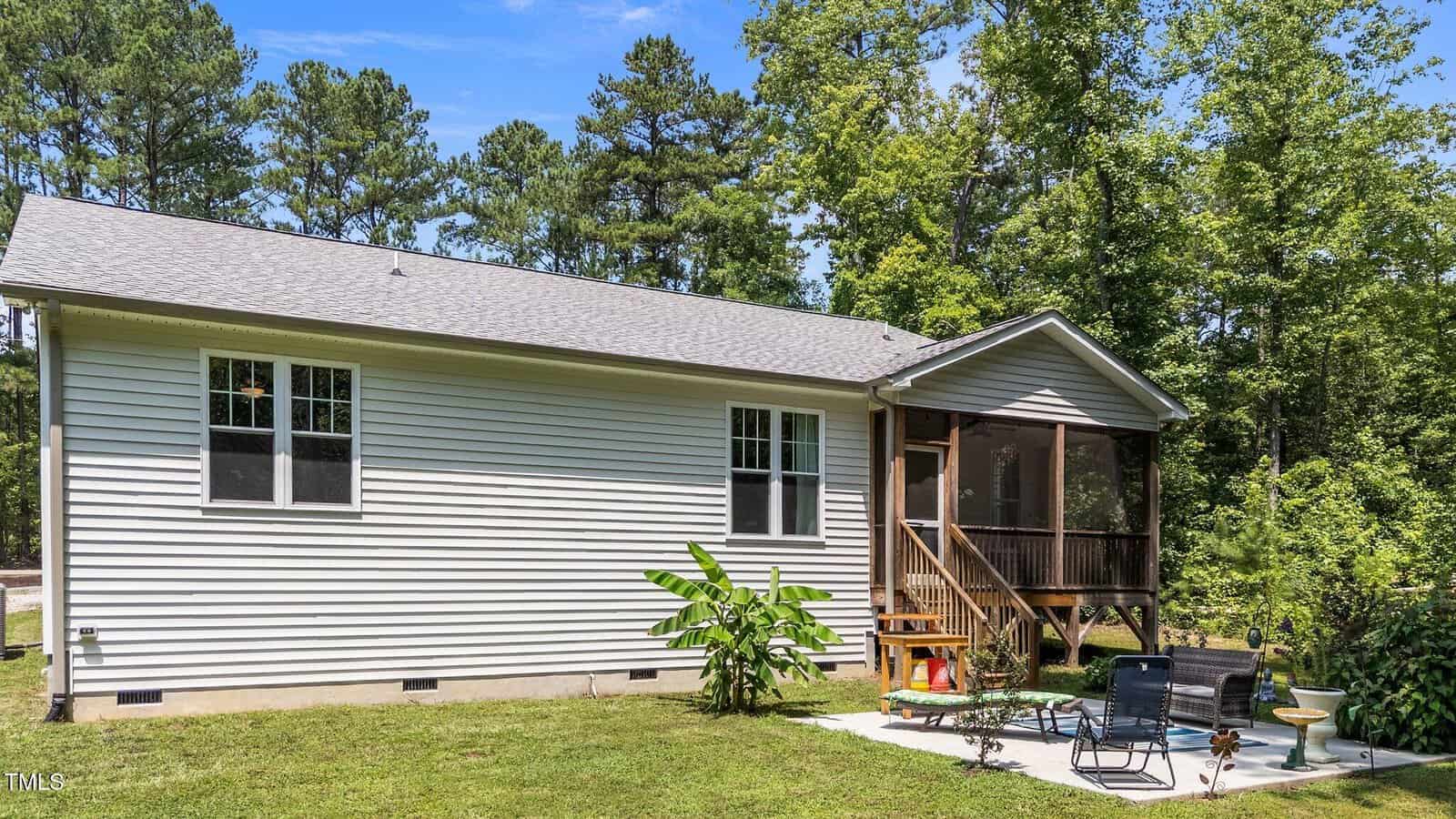 Single-story house with light-colored vinyl siding, two windows, and a screened porch at the back. There’s a small patio with outdoor chairs, a table, potted plants, and a grassy lawn, surrounded by tall trees and greenery.