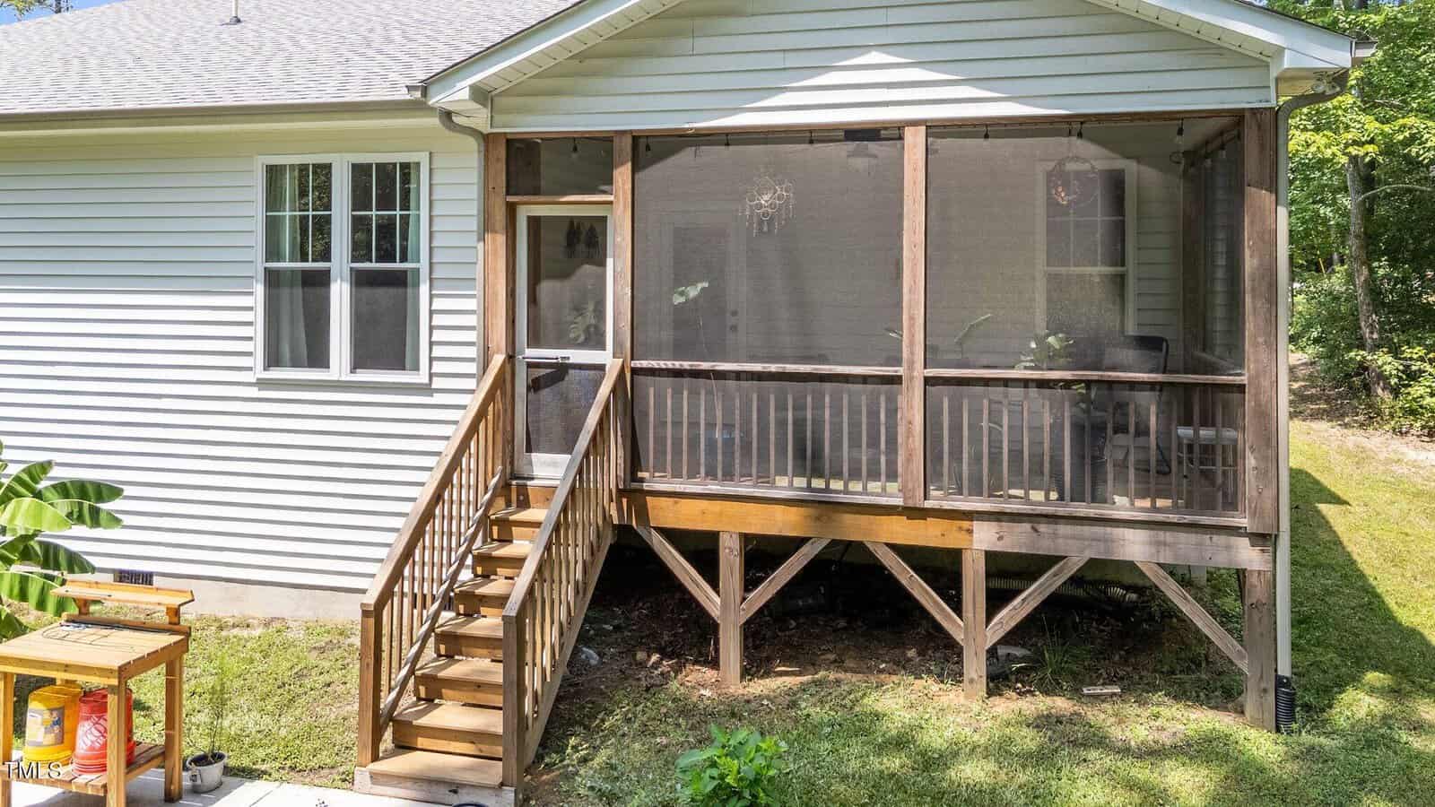A screened-in porch with wooden stairs leads up to the back entrance of a white house. The porch is elevated, supported by wooden posts, and overlooks a grassy yard with trees in the background.