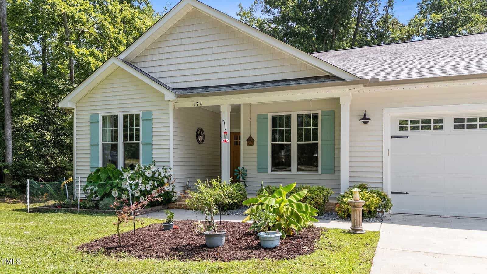 A single-story house with light-colored siding, green shutters, a front porch, potted plants, garden beds, and a driveway leading to an attached garage, surrounded by greenery and trees.