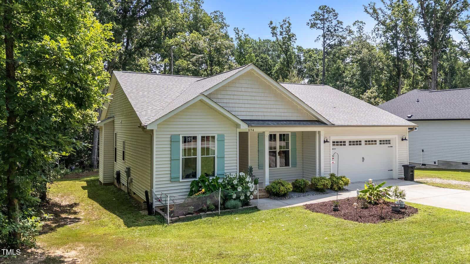 Single-story light-colored house with a covered front porch, attached two-car garage, and well-maintained lawn with shrubs and plants. Surrounded by trees and neighboring homes.