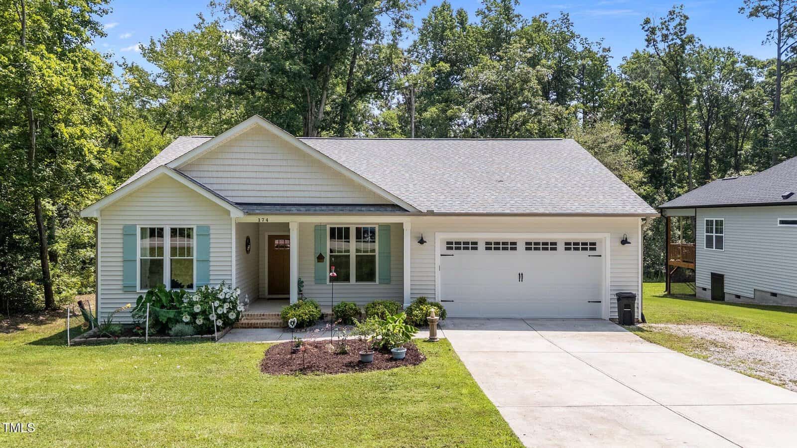 A single-story house with light blue siding, white trim, and a two-car garage. The front yard has a small garden bed, green lawn, and trees in the background. A covered porch sits by the entrance.