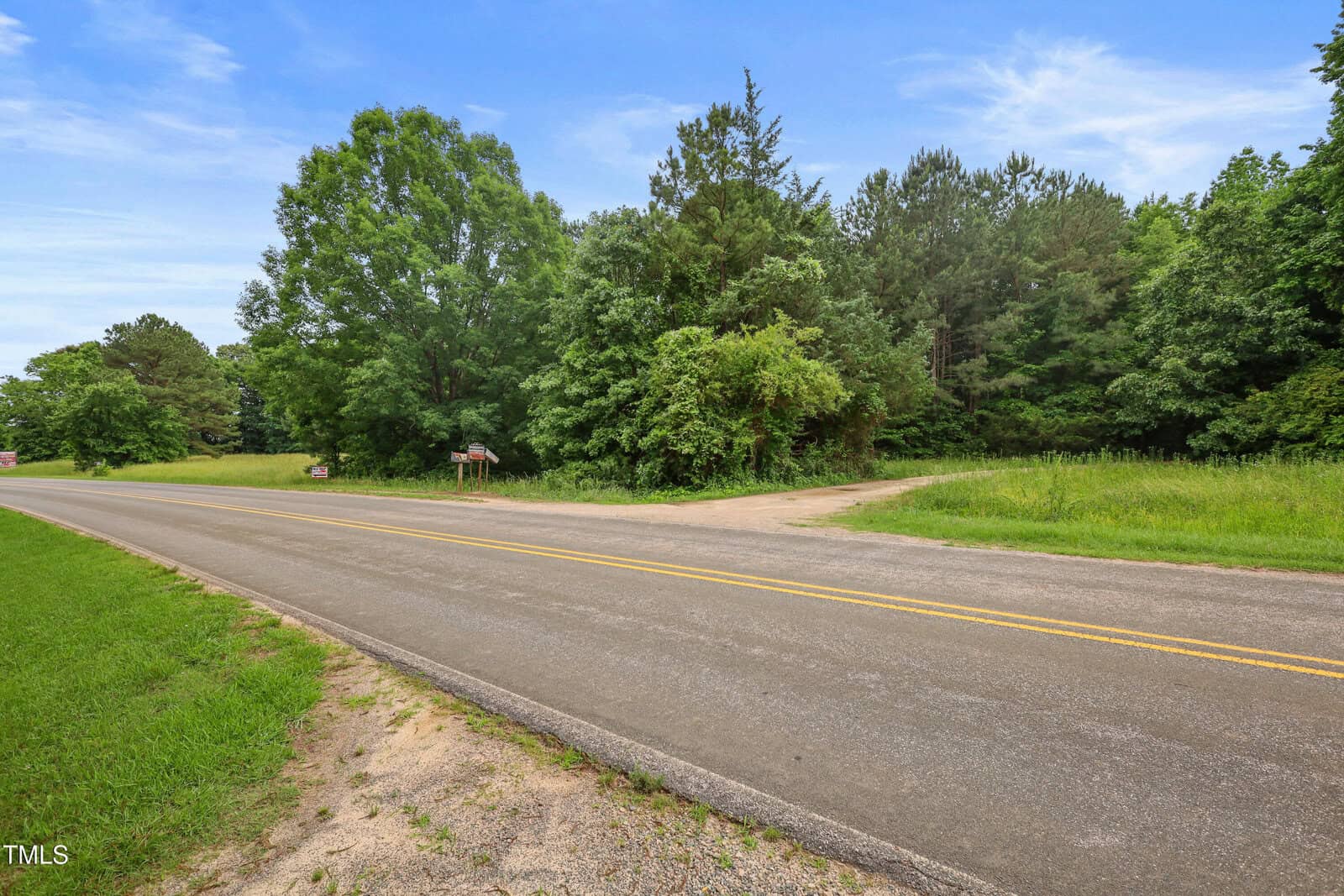 A paved two-lane road runs past a junction with a dirt road, bordered by grass and dense green trees under a partly cloudy sky.