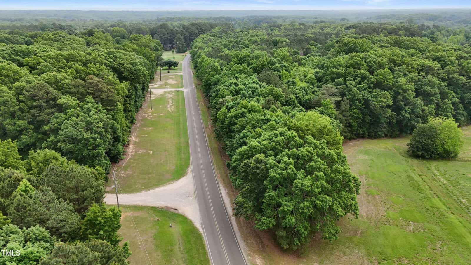A rural two-lane road runs through a green, tree-filled landscape with grass and open areas on both sides under a partly cloudy sky. Power lines line parts of the road, and the area appears calm and undeveloped.