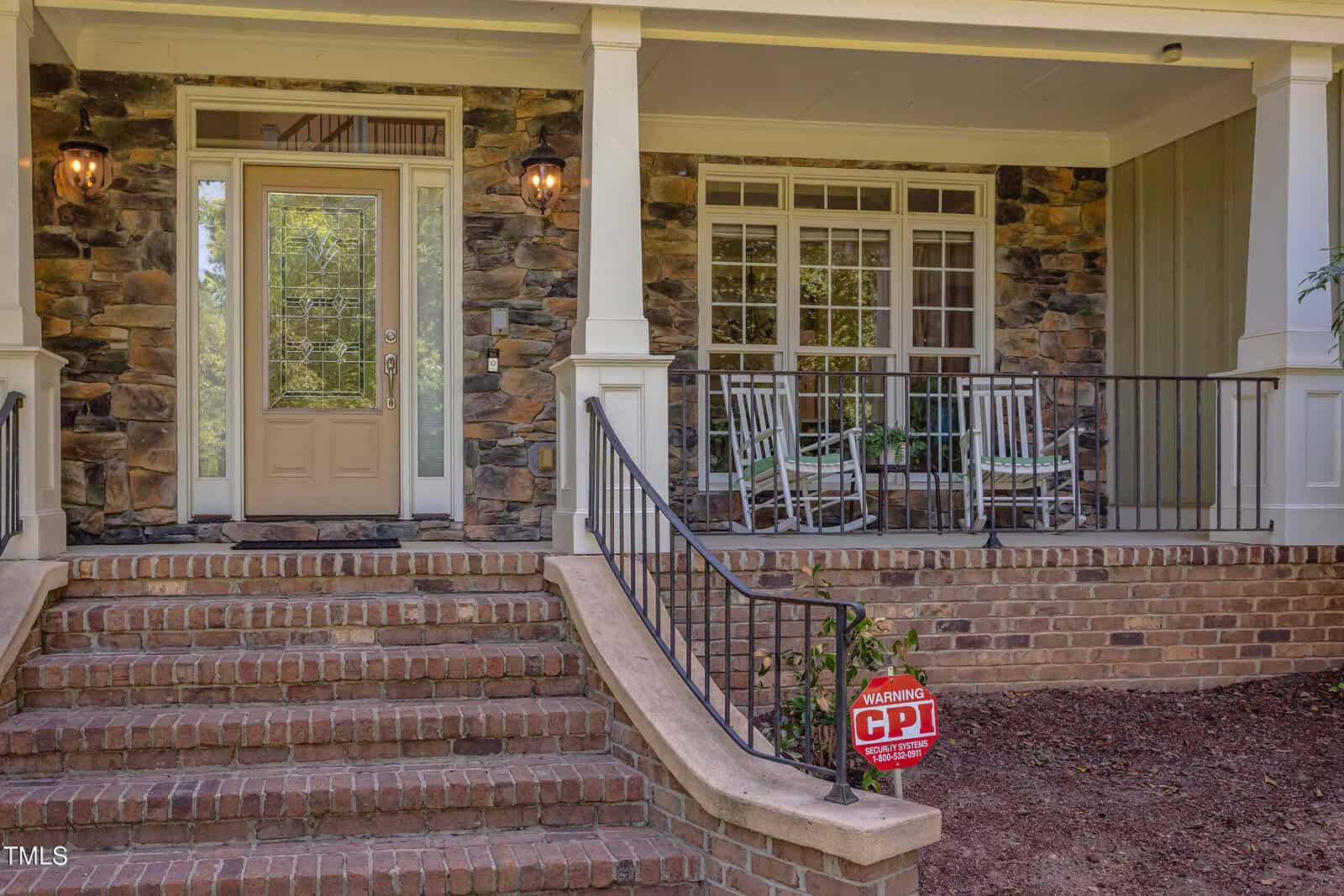 Front porch of a house with stone walls, a glass-paneled front door, two white rocking chairs, black metal railing, brick steps, and a red security sign reading "CPI." Lit lantern-style lights by the door.