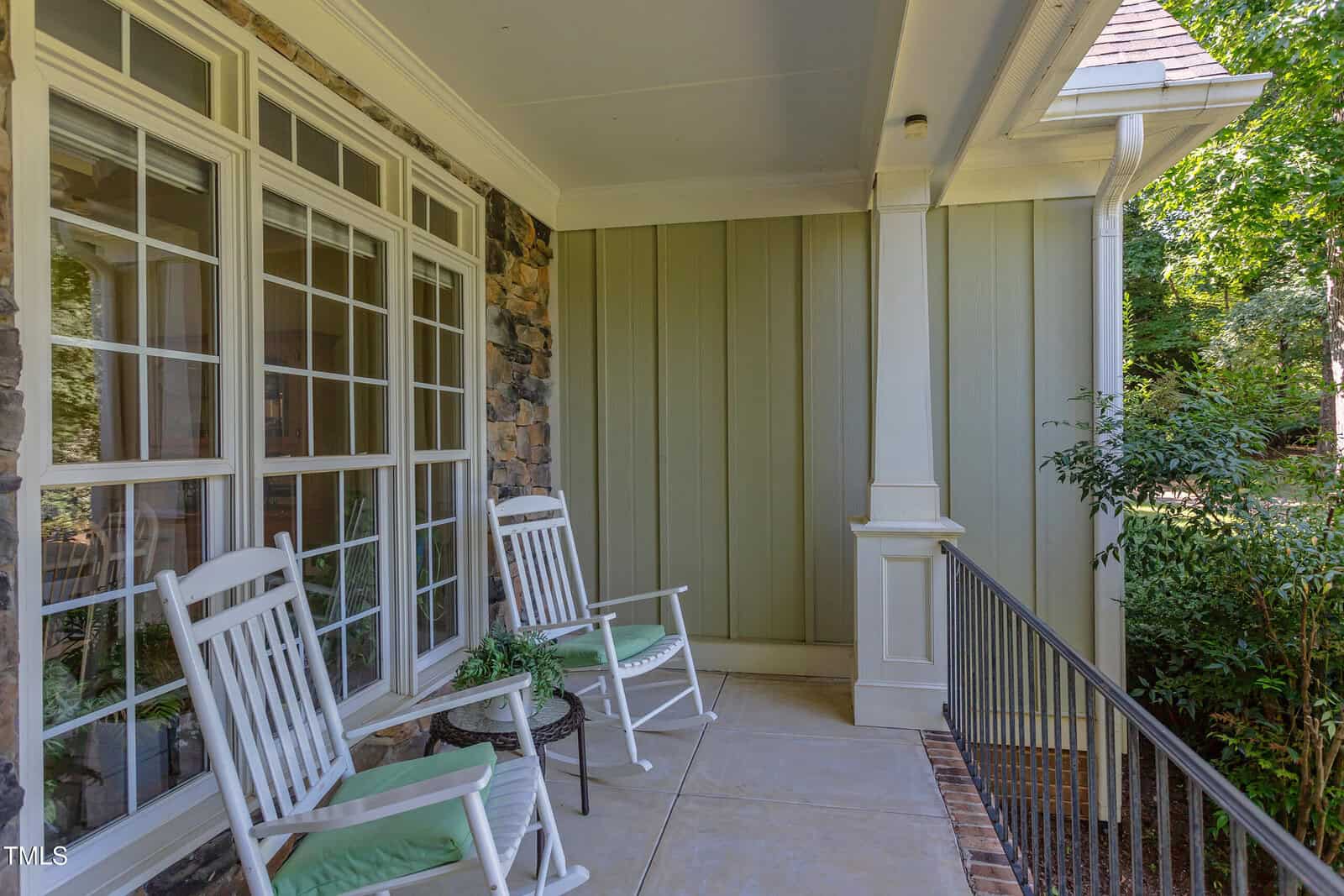 A shaded front porch with two white rocking chairs, a small table with a potted plant, large windows, stone and paneled siding, and a metal railing overlooking green plants.
