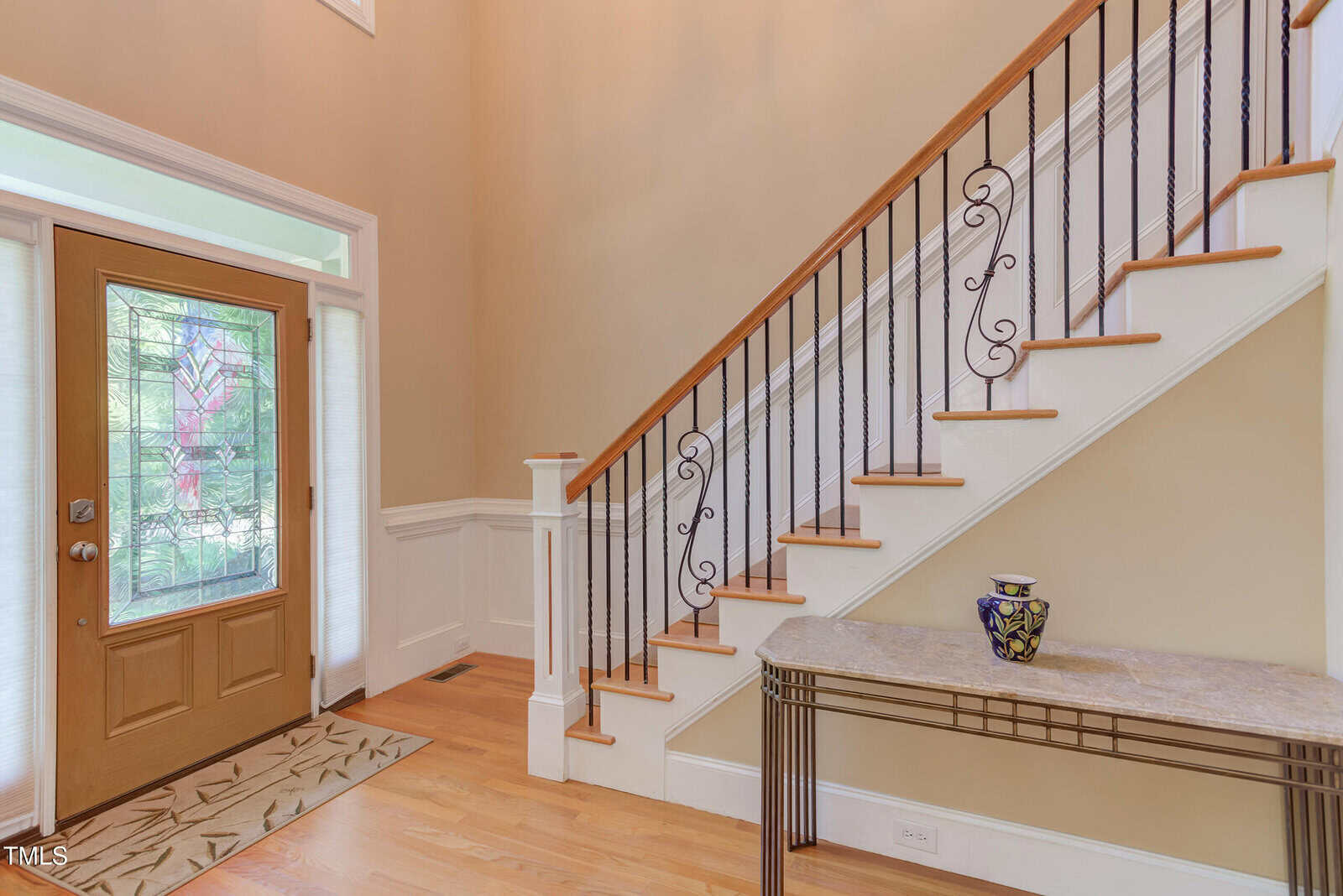 A foyer with a wooden front door featuring decorative glass, a staircase with wooden steps and black metal railings, and a console table with a patterned vase under the stairs.