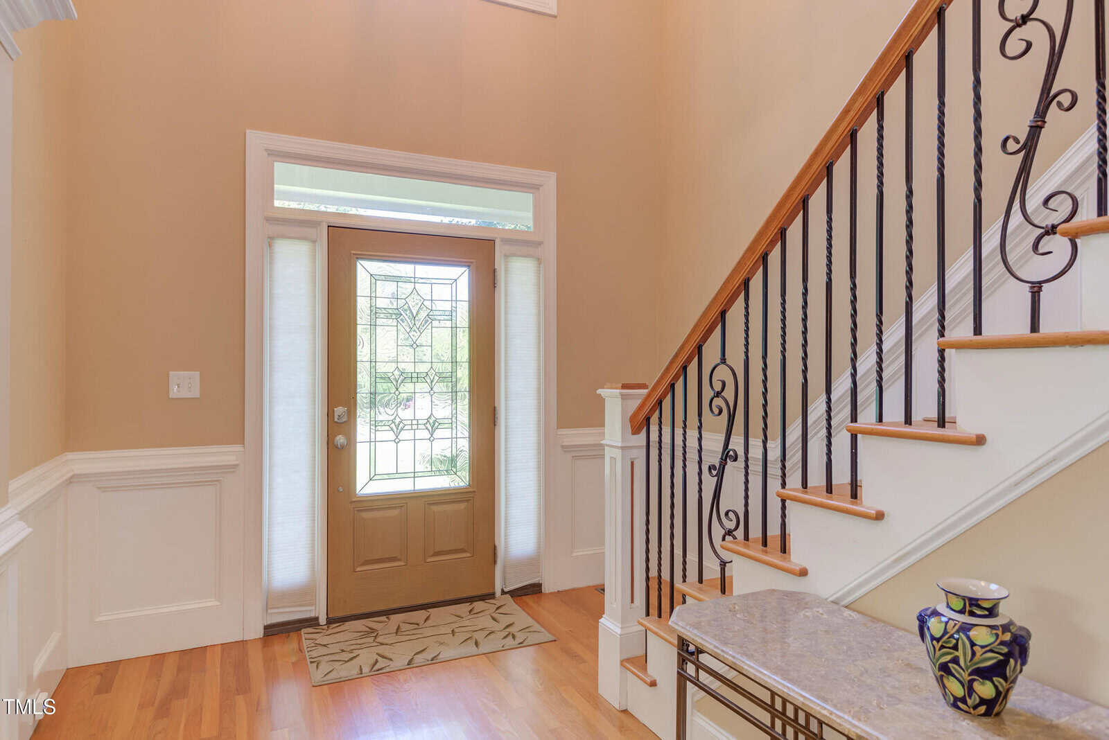 A foyer with light wood floors, a brown front door with decorative glass, white wainscoting, a staircase with wooden handrail and black iron balusters, and a table with a ceramic vase.