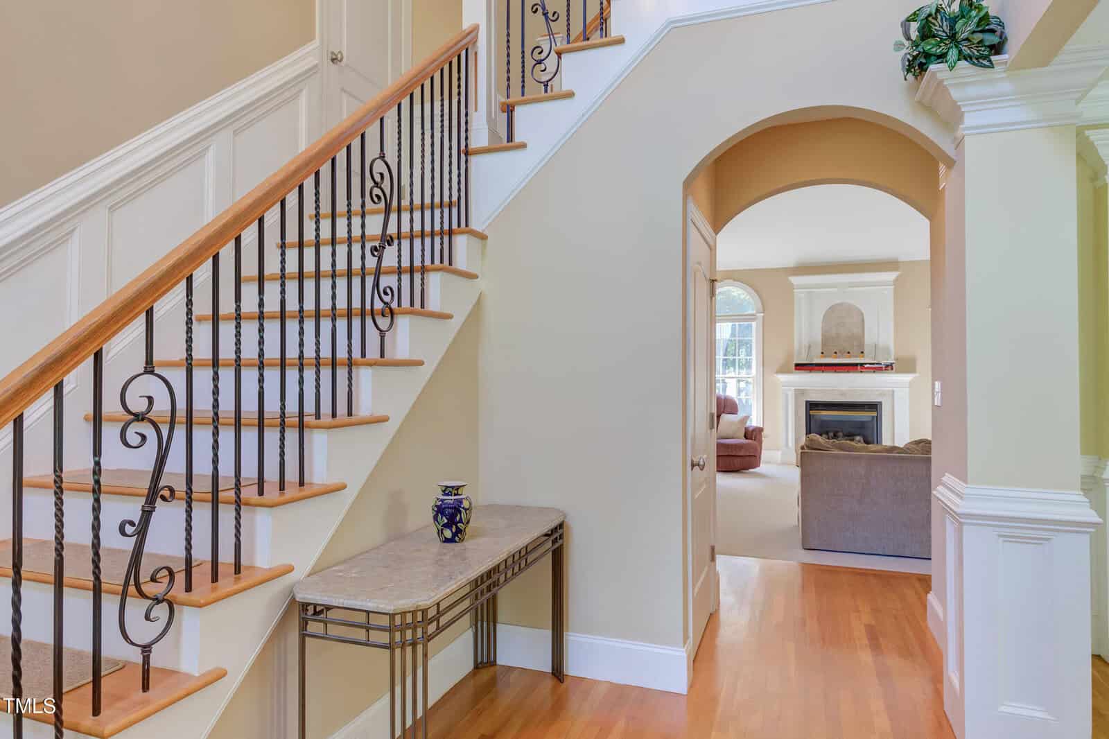 A staircase with wooden steps and decorative black iron railings next to a narrow table with a vase; an archway leads to a living room with a fireplace and sofas.